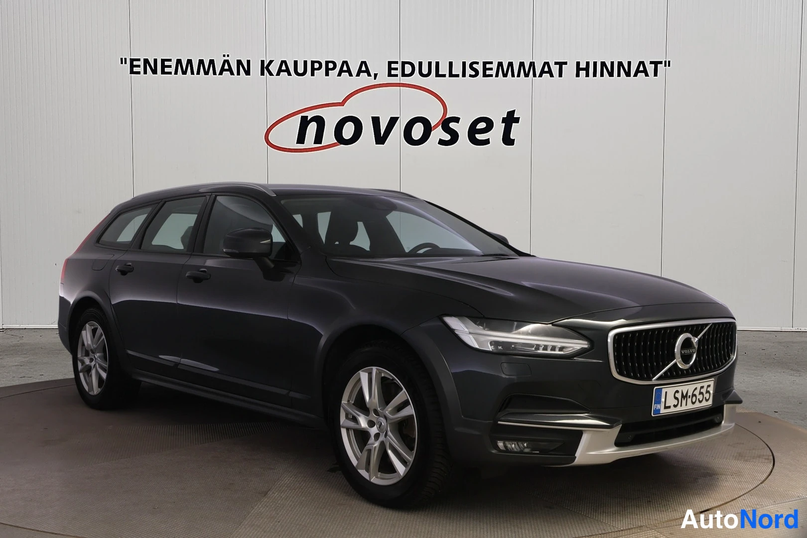 Etuviisto, nokka oikealle: Musta Volvo V90 Cross Country -auto esillä. Etuosa ja oikea sivu näkyvät, jossa on kromipaneelit etupuskurissa ja takatolkussa. Auton renkaisiin on asenn