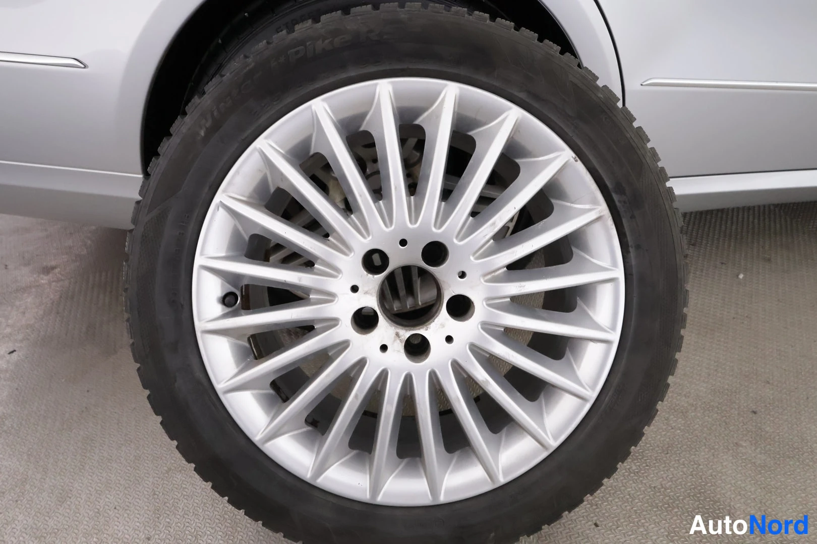 Sivulta: Lähikuva auton pyörästä: hopeanvärinen monisakarainen vanne mustalla, uritetulla renkaalla; auton vaalean hopeanvärinen kylki näkyy kuvan reunoilla.