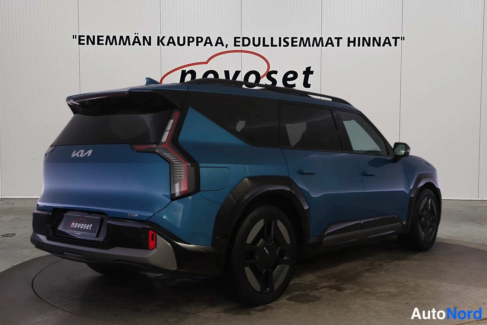 Takaviisto: Sinivihreä Kia EV9 takaviistosta kuvattuna valkoisella studiotaussilla, perä vasemmalla; kattokaiteet ja tummat vanteet näkyvät.