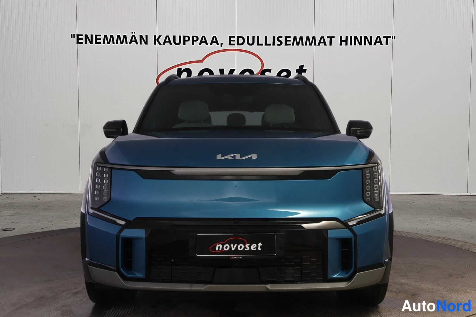 Edestä: Sininen henkilöauto suoraan edestä päin kuvattuna, KIA-merkki konepellissä, LED-ajovalot ja novoset-tekstillä varustettu rekisterikilven paikka näkyvissä, taust