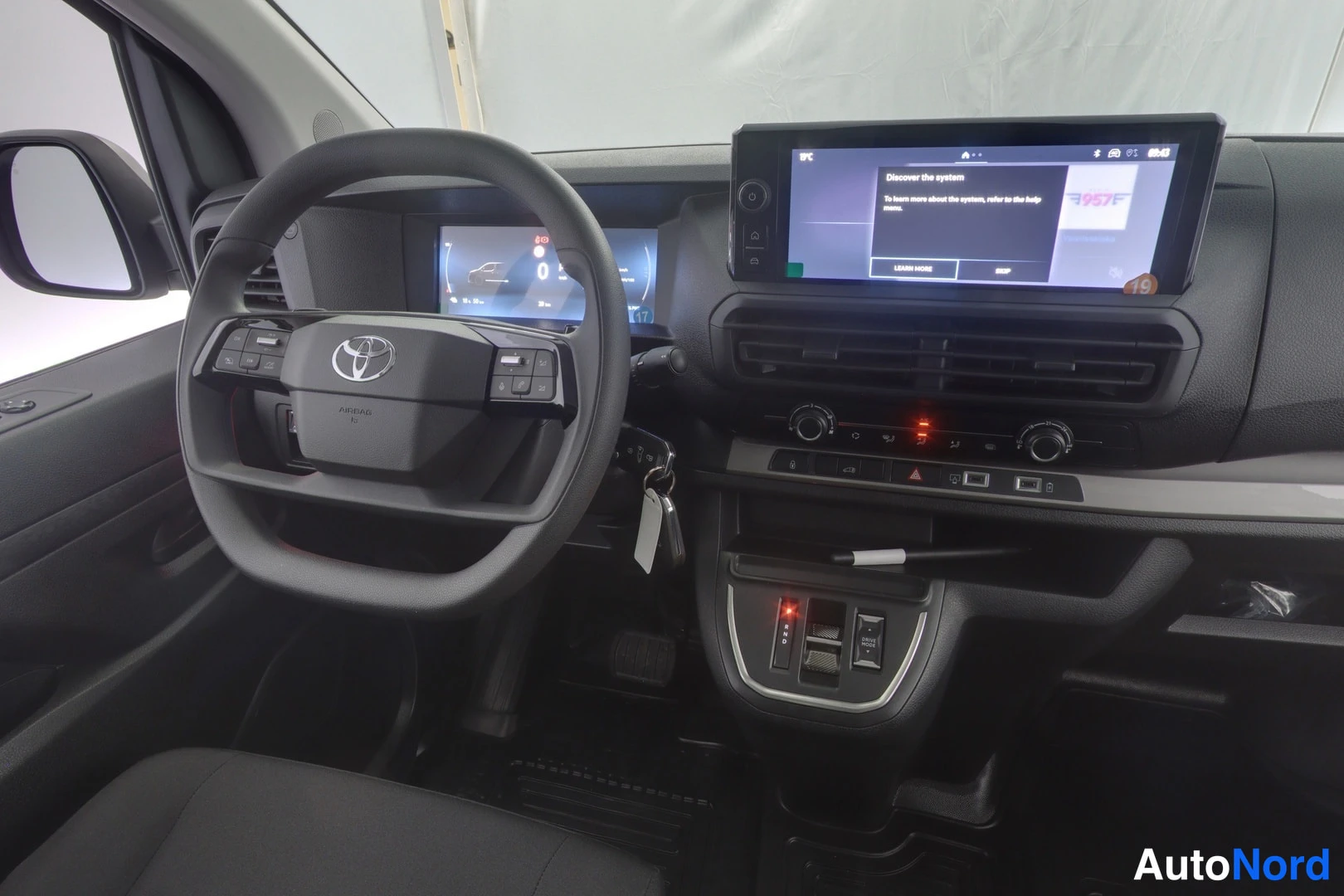 Muu: Lähikuva ohjaamosta: Toyota-ohjauspyörä, mittariston näyttö ja keskusnäyttö, jossa teksti 'Discover the system'.