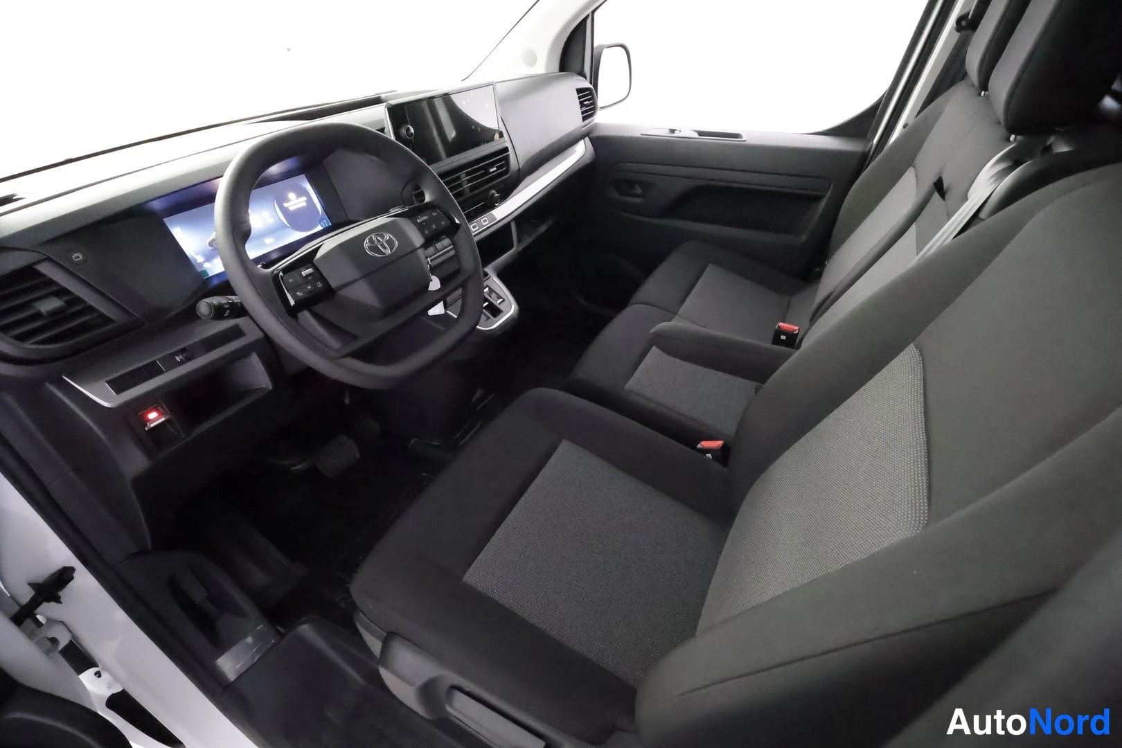 Etuistuimet: Front interior view from open passenger door showing white body frame. Dashboard has a touchscreen display. Dark grey fabric front seats are visible. A red illu