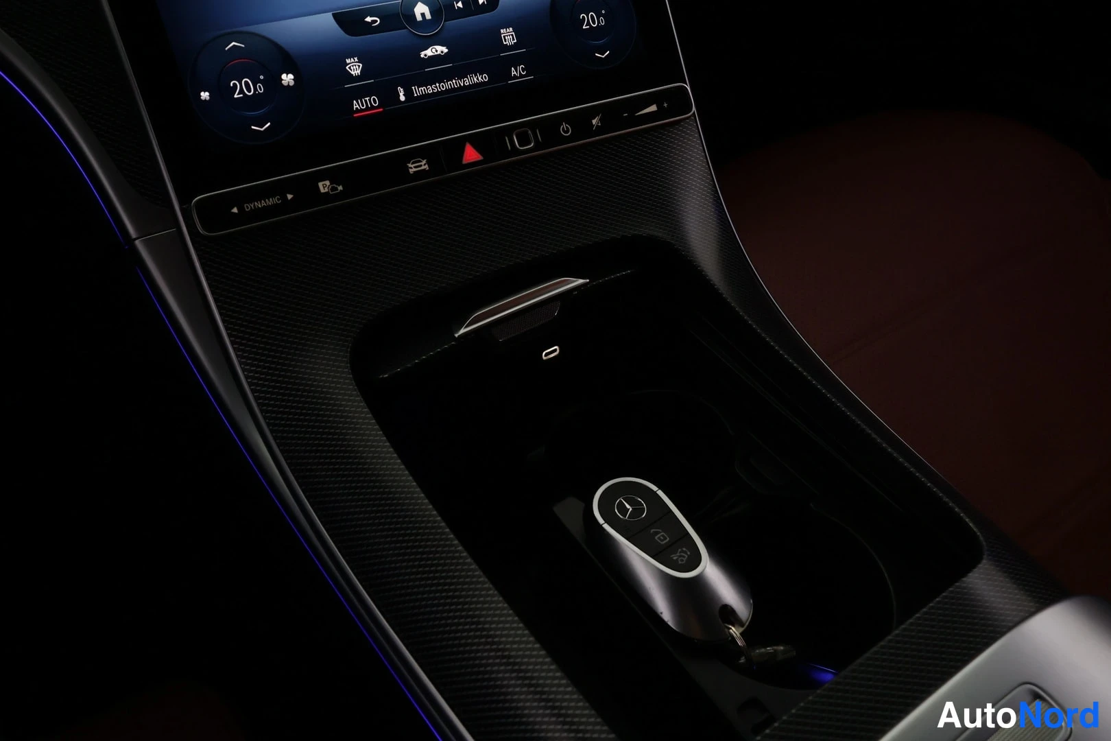 Muu: Keskikonsolin lähikuva: kosketusnäyttö ilmastointivalikolla (20,0°, AUTO, A/C), DYNAMIC-valitsin ja Mercedes-avain konsolin lokeroon sijoitettuna, sininen koros
