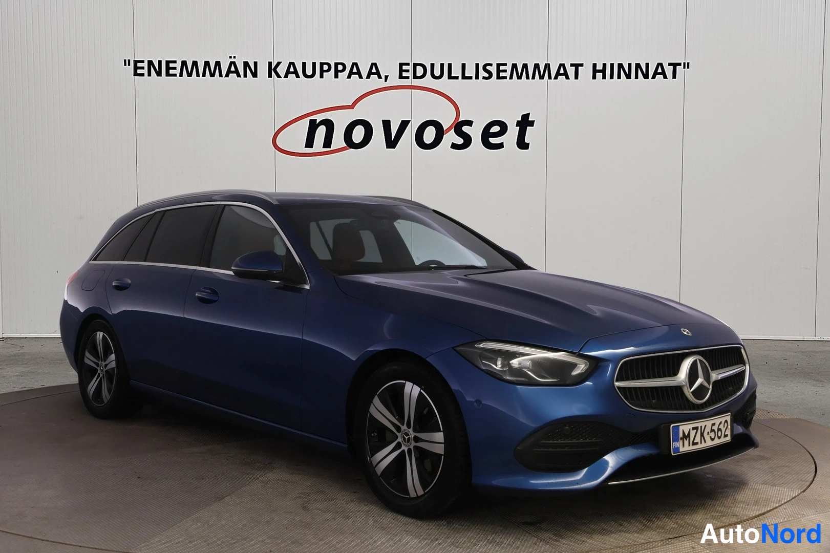 Etuviisto, nokka oikealle: A blue station wagon shown from a front three-quarter angle with the grille positioned to the right of the rear wheel on the vehicle side. Features alloy