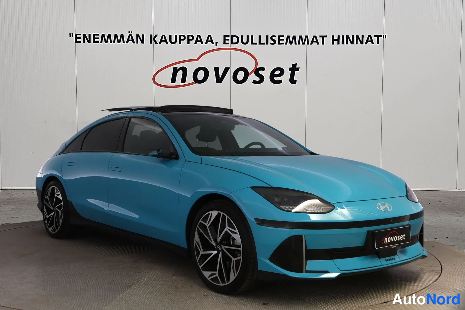 Etuviisto, nokka oikealle: Turkoosinsininen Hyundai IONIQ 6 etukulmakuvassa nokka oikealla, valkoista showroom-seinää vasten; takapyörä ja ovet näkyvät vasemmalla.