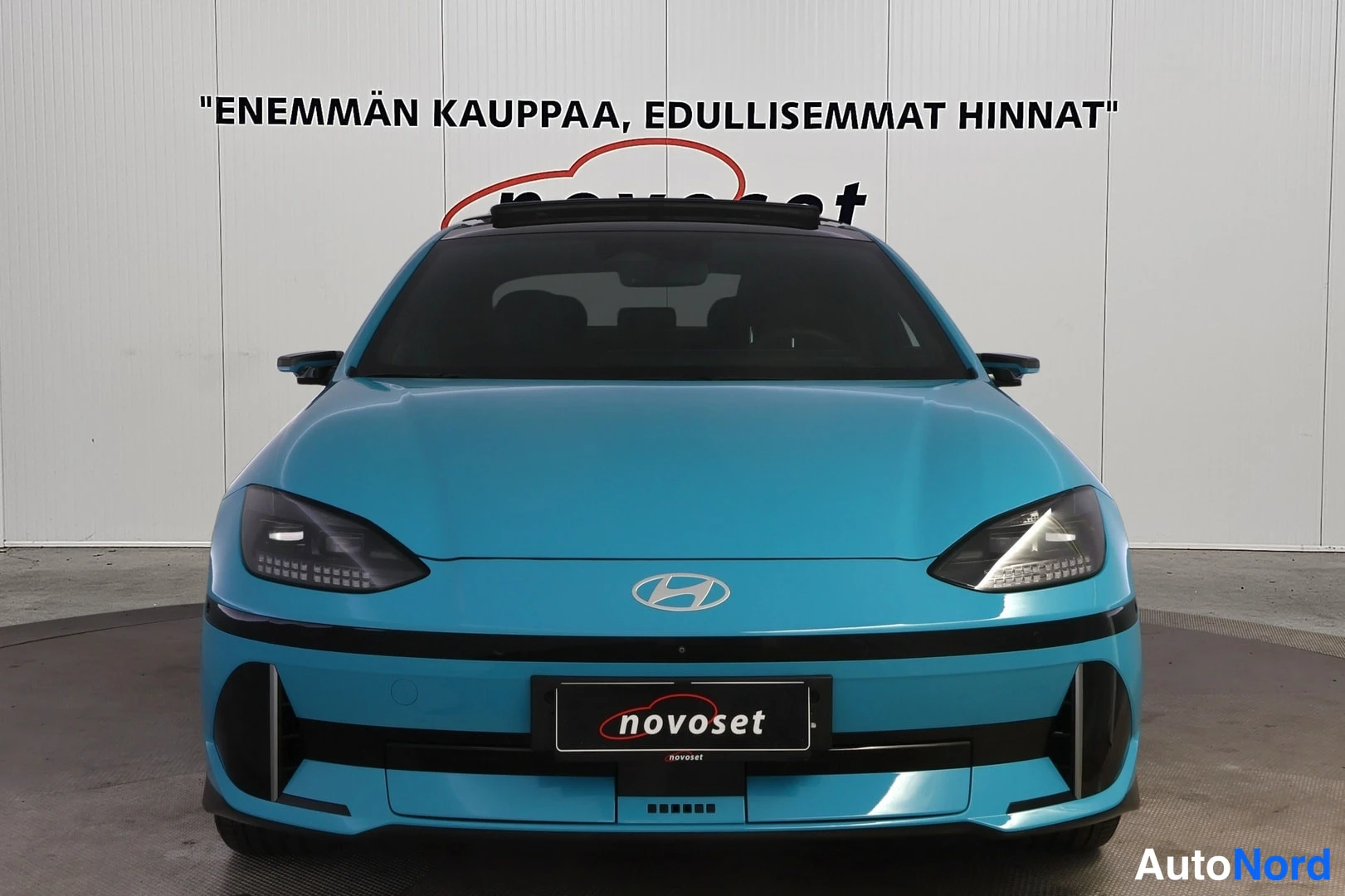 Edestä: Hyundai IONIQ 6 suoraan edestä kuvattuna; Hyundai-merkki ohuine ajovaloineen ja novoset-kilpi etupuskurissa, valkoinen tausta.