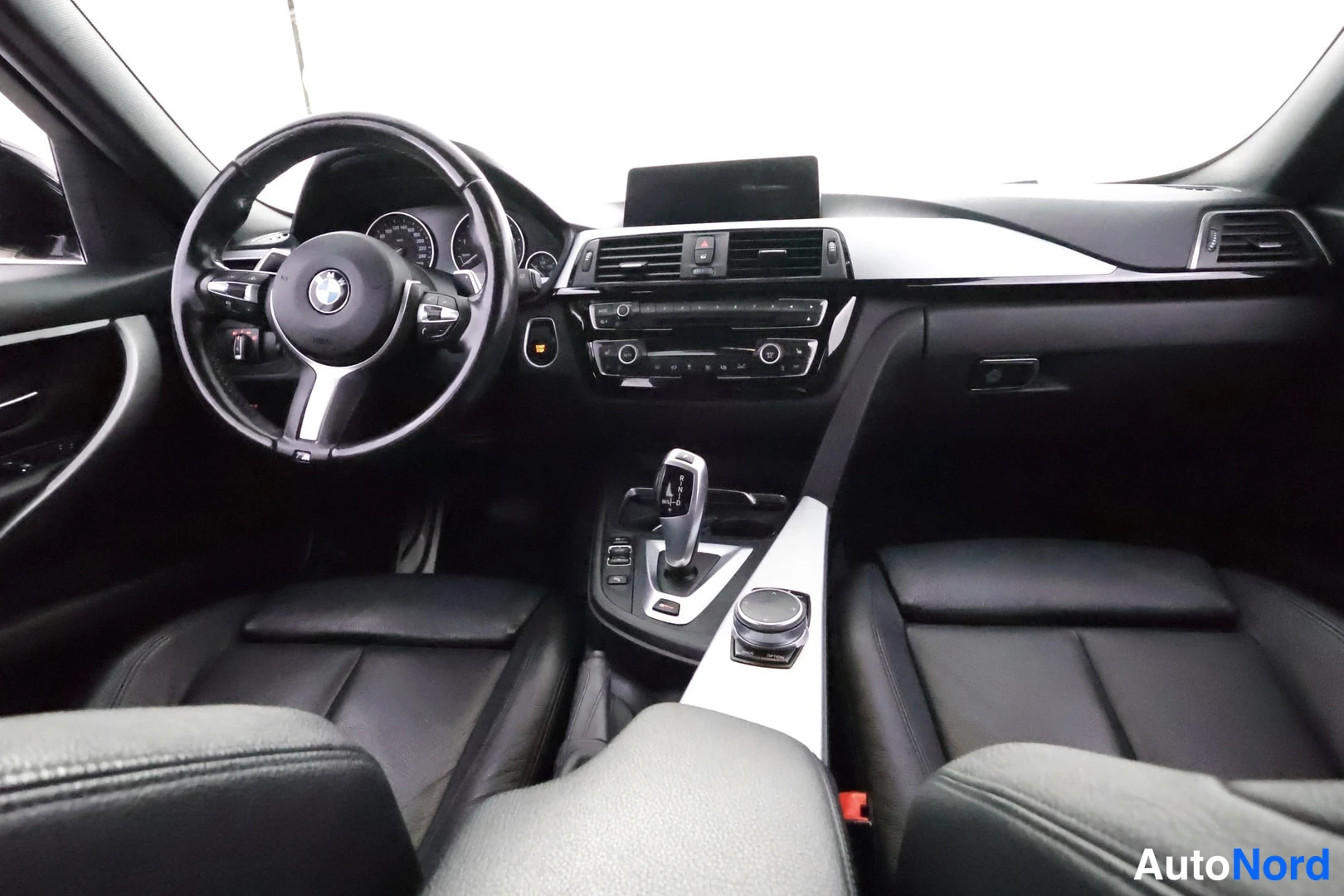 Muu: Sisätila kuljettajan puolelta kuvattuna: BMW-ohjauspyörä, mittaristo, automaattivaihteen valitsin ja kierto-ohjain keskikonsolissa, mustat etuistuimet.