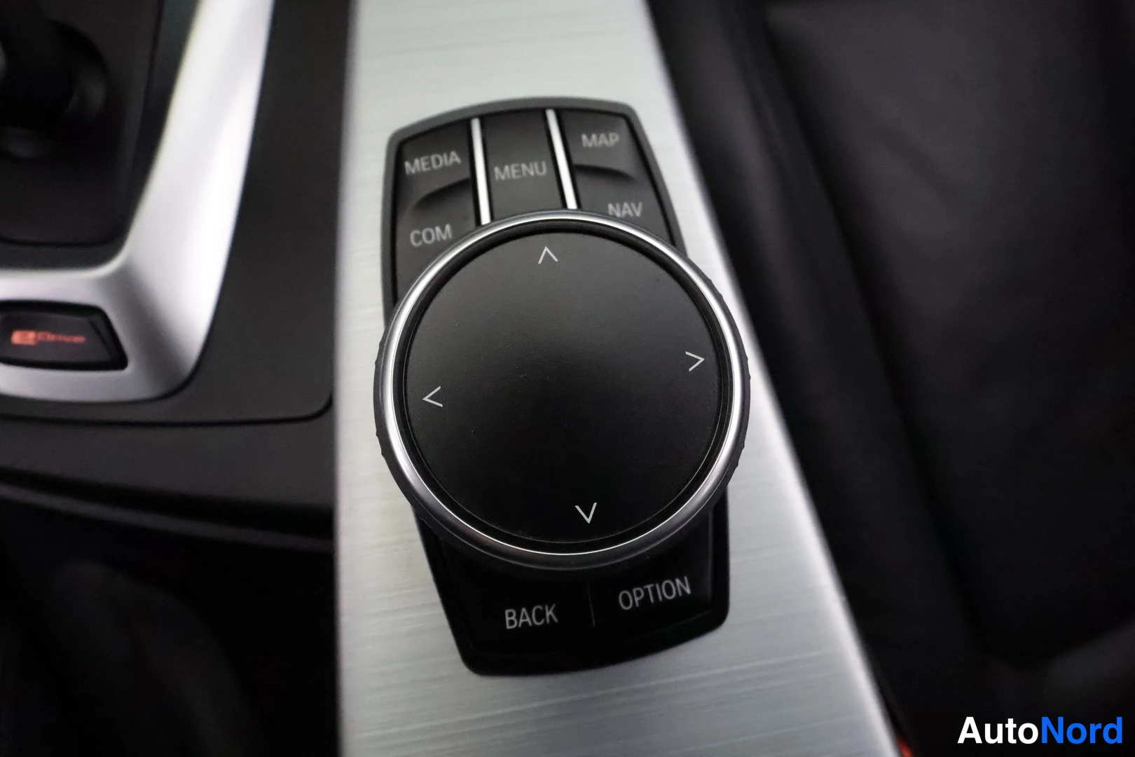 Yksityiskohta: Sisätilakuva iDrive-ohjaimesta: pyöreä ohjainnäppäin suuntanuolilla, ympärillä MEDIA, MENU, MAP, NAV, COM, BACK ja OPTION -painikkeet, harmaa alumiinikoriste.