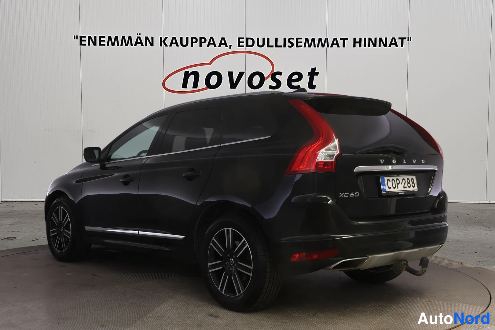 Takaviisto: Musta Volvo XC60 takaviistosta vasemmasta kulmasta, rekisteritunnus, valkoisen Novoset-mainosseinän edessä.