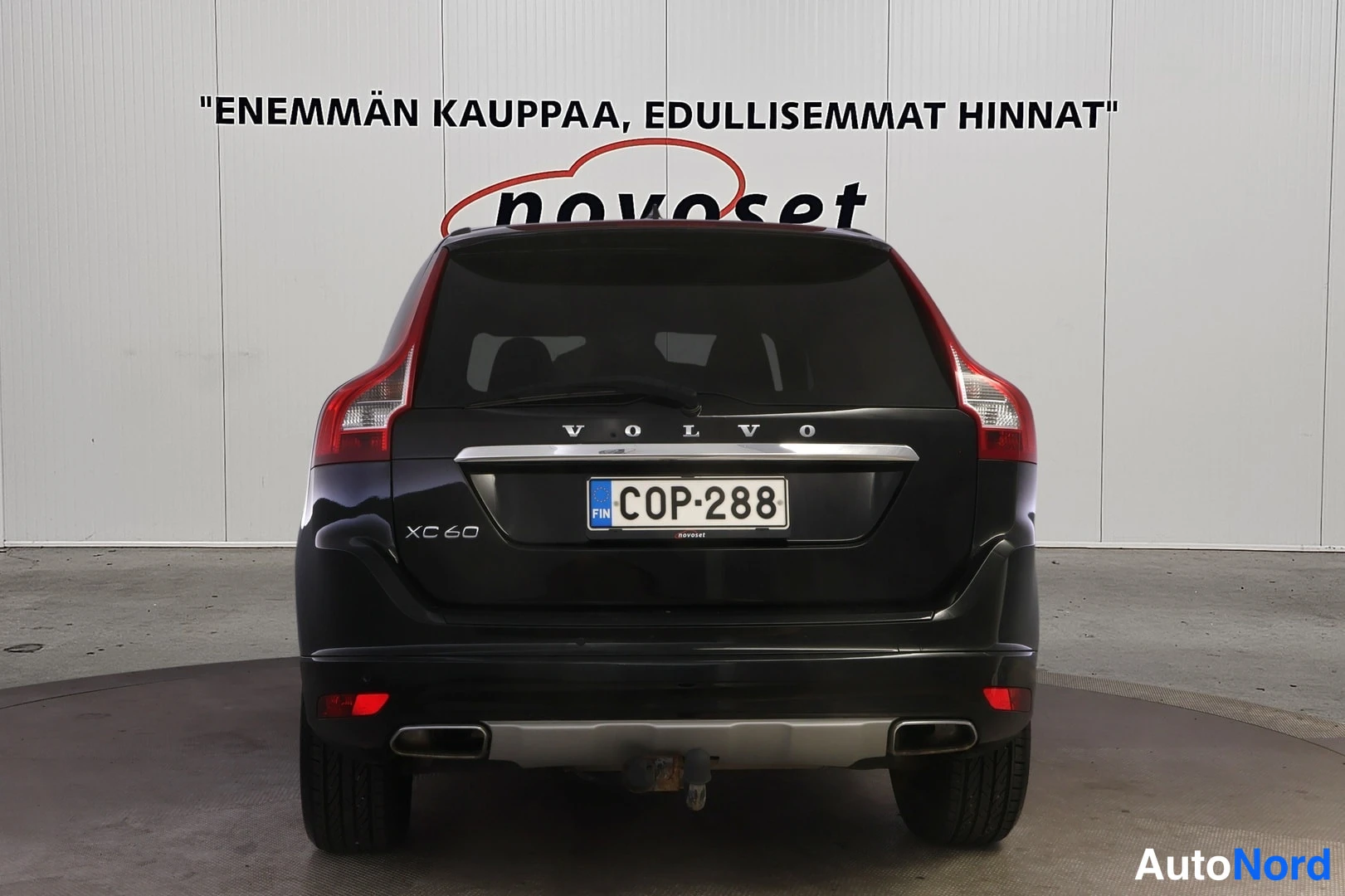Takaa: Musta Volvo XC60 suoraan takaa kuvattuna, rekisteritunnus, valkoisen Novoset-mainosseinän edessä.