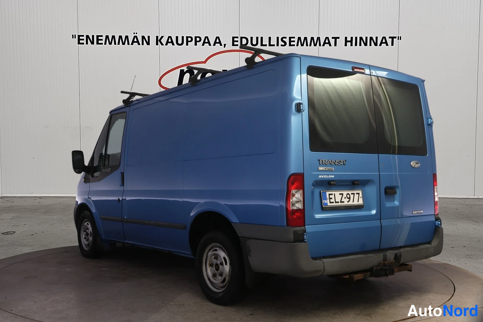 Takaviisto: Sininen pakettiauto takaviistossa oikealta, Transit-tunnus ja kattotelineet.