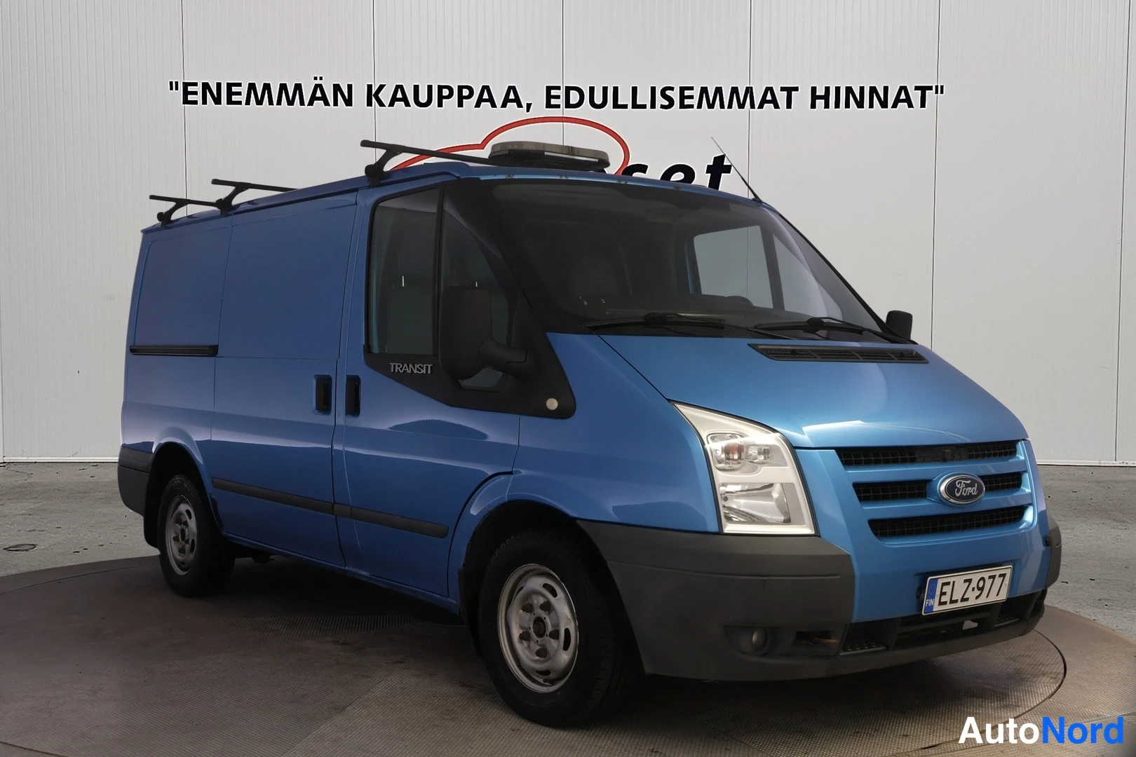 Etuviisto, nokka oikealle: Sininen pakettiauto kolmivälinäkymässä edestä oikealta, kattoteline kiinnitettynä, rekisteritunnus.
