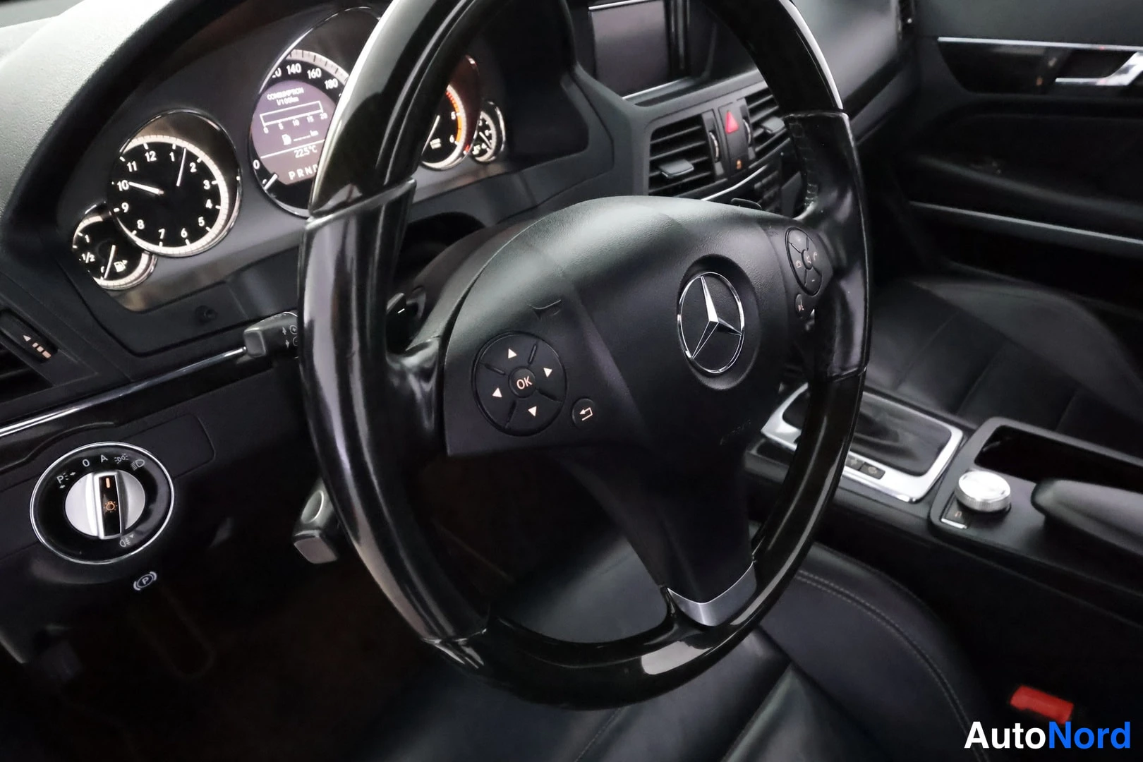 Edestä: Mercedes-Benzin ohjaamo: musta rakenneohjaurointi ja merkkivalot sekä mittaristo, jossa näkyy kellonaika, lämpötila 23,5 °C ja vaihdetila P.