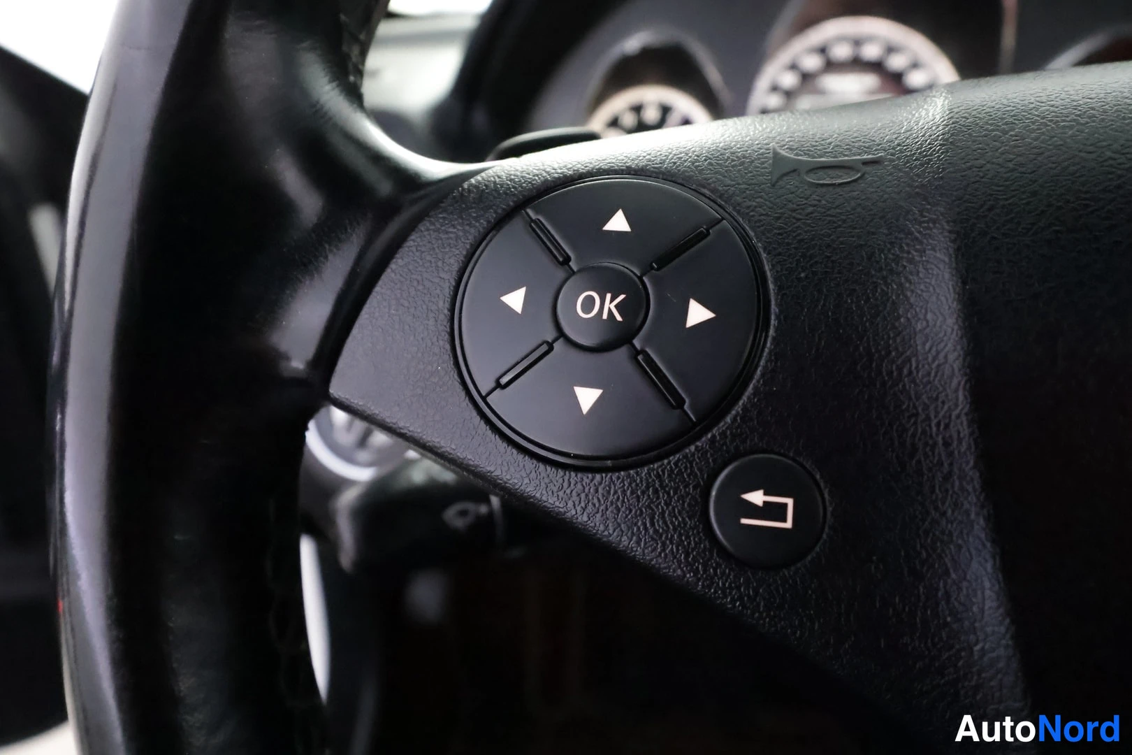 Edestä: Lähikuva Mercedes-Benzin ohjauspyörän vasemmasta painikeryhmästä: nuolinäppäimet ja OK-painike, alapuolella palautuspainike.