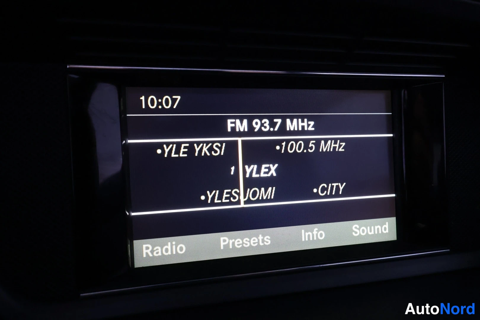 Edestä: Auton radio-näyttö kuvaajalla FM 93.7 MHz ja kanavalista Yle-asemilla, kello 10:07.