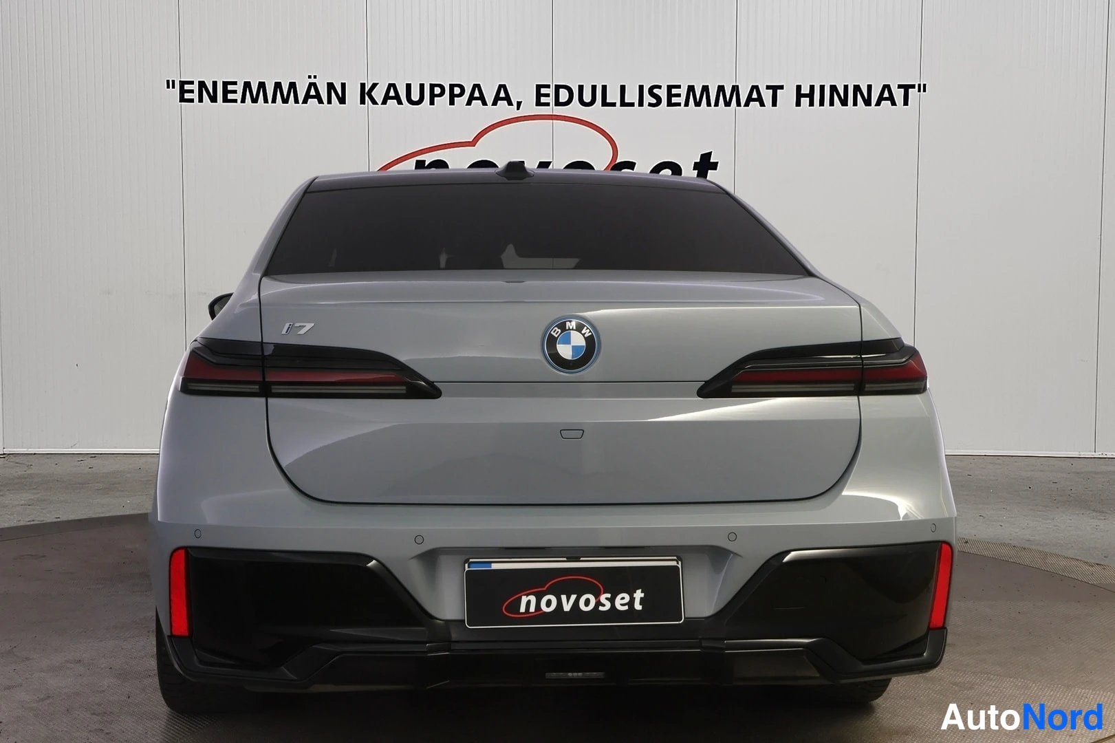 Takaa: Harmaa BMW i7 -sedan suoraan takaa: kapeat takavalot, i7-merkintä, sinivalkoinen BMW-merkki ja novoset-kilpi takapuskurin alueella.