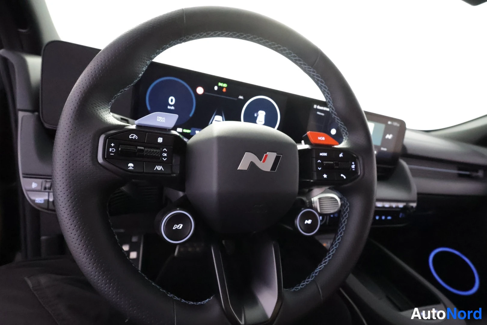 Yksityiskohta: Kokonaisnäkymä ohjauspyörästä, jossa Hyundai N -logo, molemmin puolin nappiryhmiä ja DRIVE MODE- ja NGB-vivut, taustalla kojelaudan näytöt.