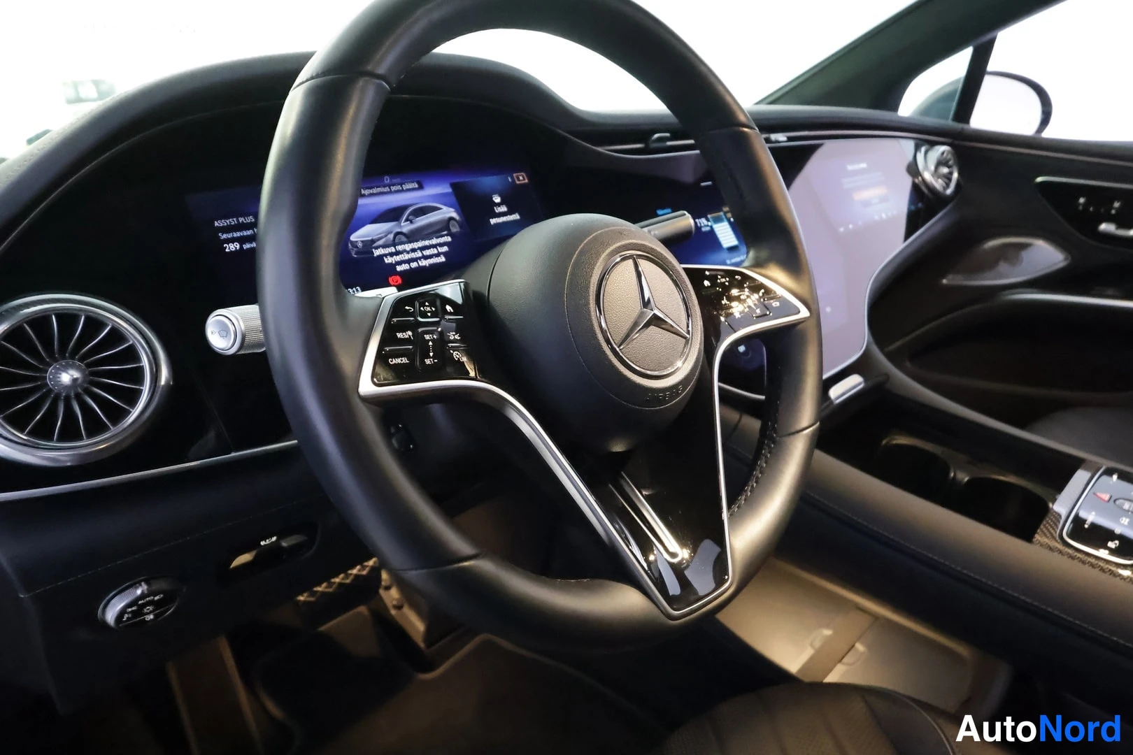 Muu: Kuljettajan näkymä Mercedes-Benzin ohjauspyörään keskuspalkilla olevaan Mercedes-tähteen; taustalla pitkä näyttöpaneeli ja ilmastointisuuttimet, kojelaudan näyt