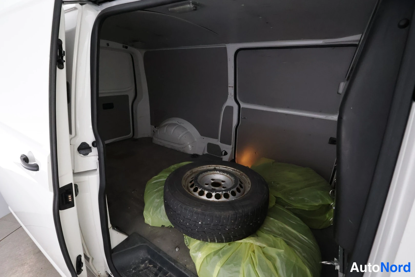 Muu: Pakettiauton kuormatila avoovista ovista: tyhjät paneeliseinät, lattialla vararengas ja vihreitä muovipusseja.