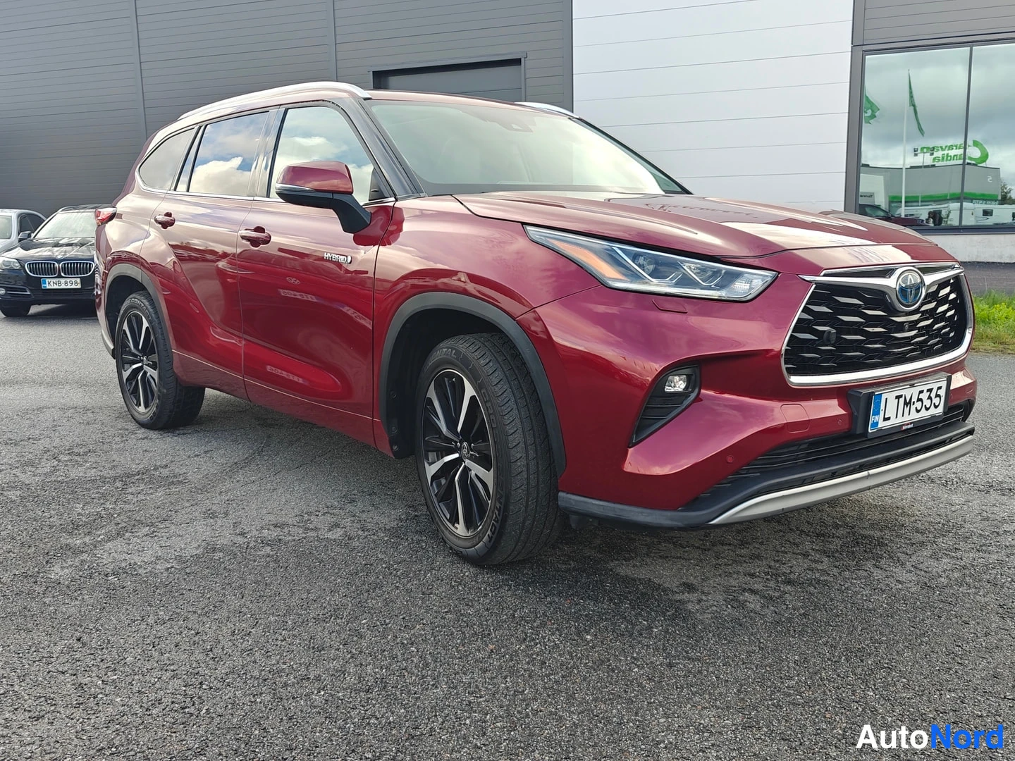 Etuviisto, nokka oikealle: Tummanpunainen Toyota Highlander kuvattuna vinosti edestä, keula oikealla. Etusäleikkö, ajovalot, rekisteritunnus ja Hybrid-merkintä etovessa näkyvät.