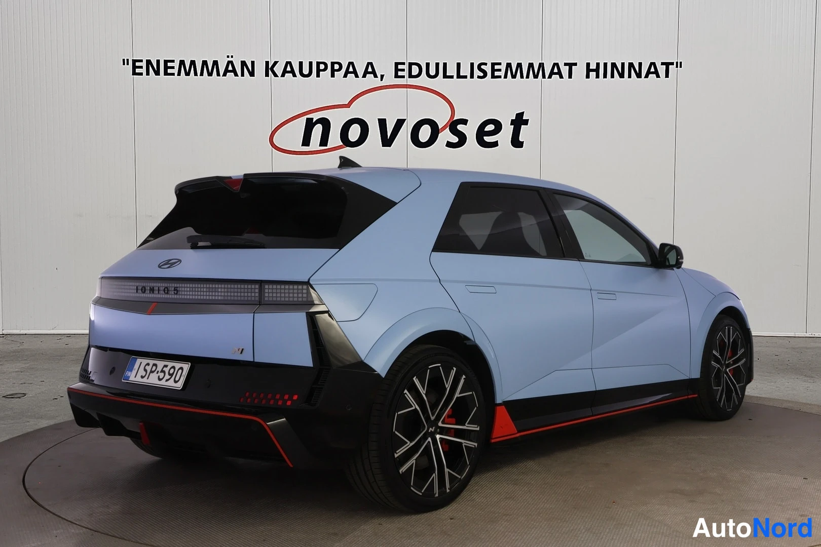 Takaviisto: Vaaleansininen auto takaviistosta oikealta, takavalo- ja kylkialue näkyvissä, mustat vanteet ja punaiset sivukorostukset.