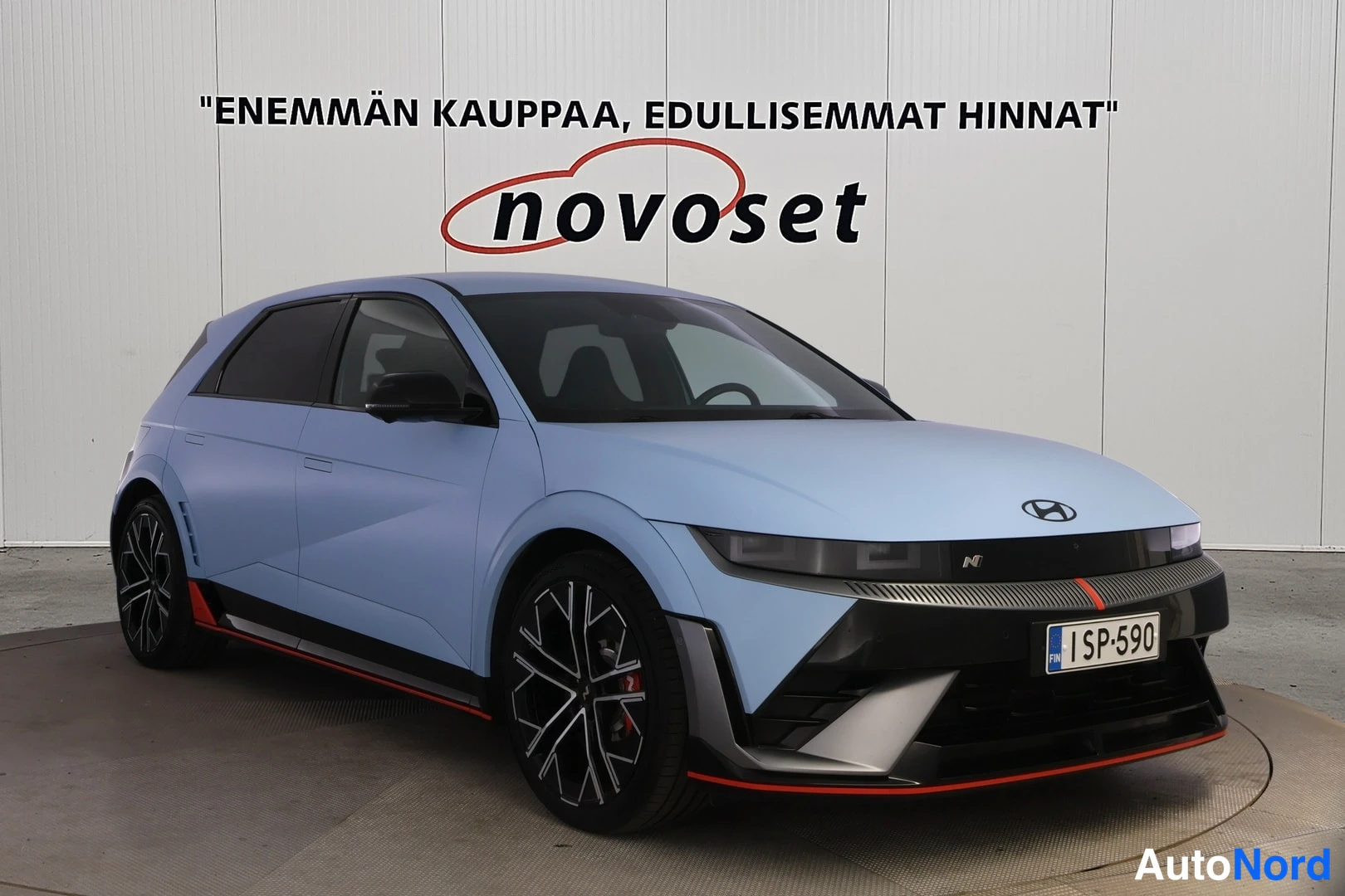 Etuviisto, nokka oikealle: Vaaleansininen Hyundai IONIQ 5 etukolmanneskulmakuvassa showroomin valkoista seinää vasten, keula osoittaa oikealle; mustat vanteet ja rekisterikilpi.