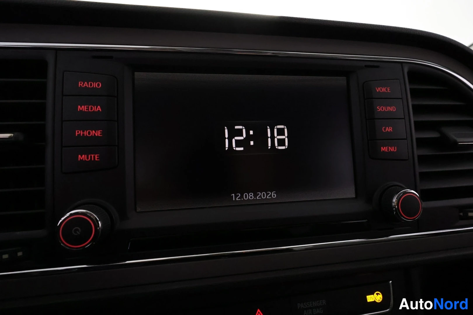 Yksityiskohta: Autoradion näytön lähikuva: kello 12:18 ja päivämäärä 12.08.2026, ympärillä RADIO-, MEDIA-, PHONE-, MUTE-, VOICE-, SOUND-, CAR- ja MENU-napit.