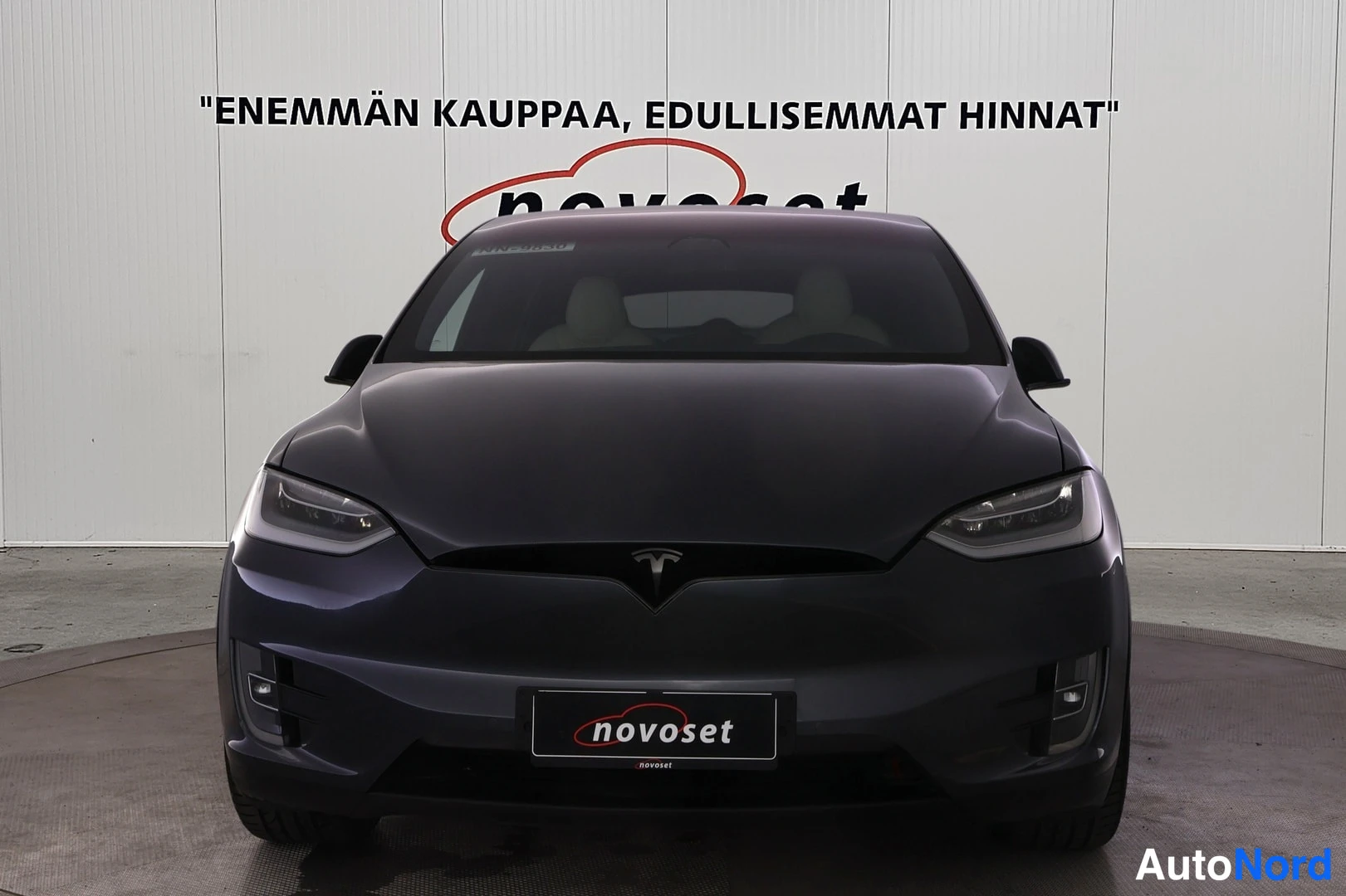 Edestä: Tesla Model X suoraan edestä; tumma kori, ohuet LED-ajovalot ja novoset-kyltti etupuskurin paikalla.