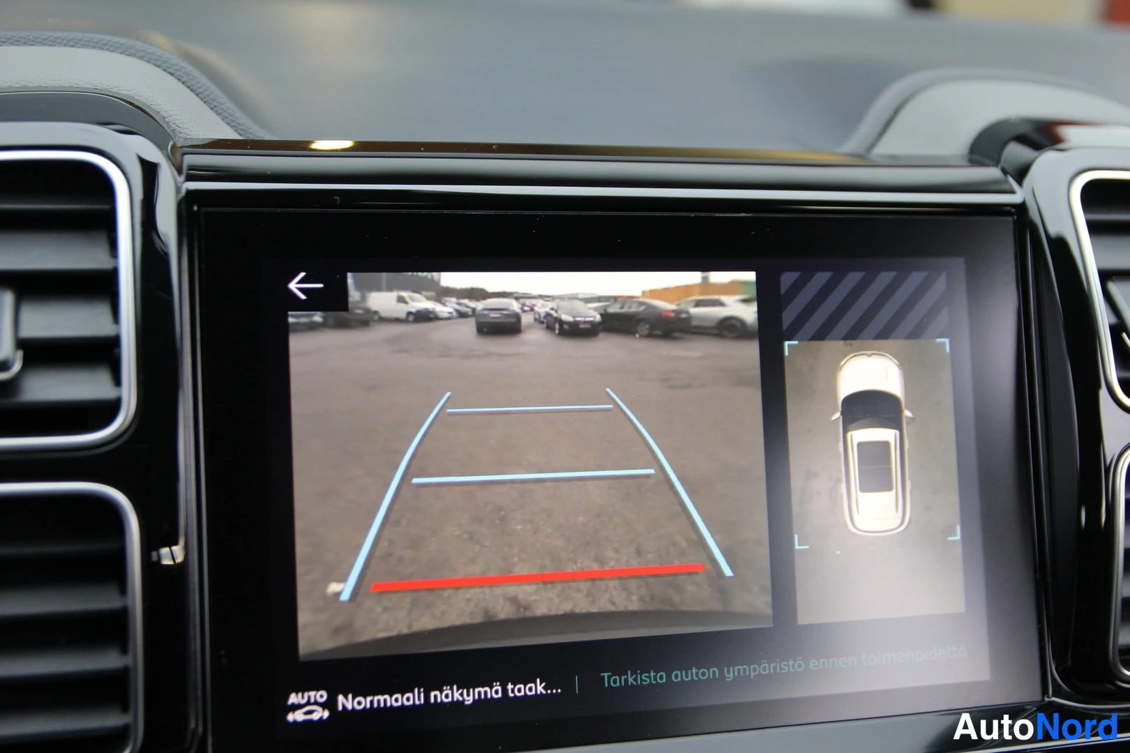 Yksityiskohta: Infotainment-näyttö, jolla näkyy peruutuskameran kuva sinisine kaartuine opastusviivoineen ja punaisine etäisyysviivoineen sekä ajoneuvon ylhäältä nähty kuva.