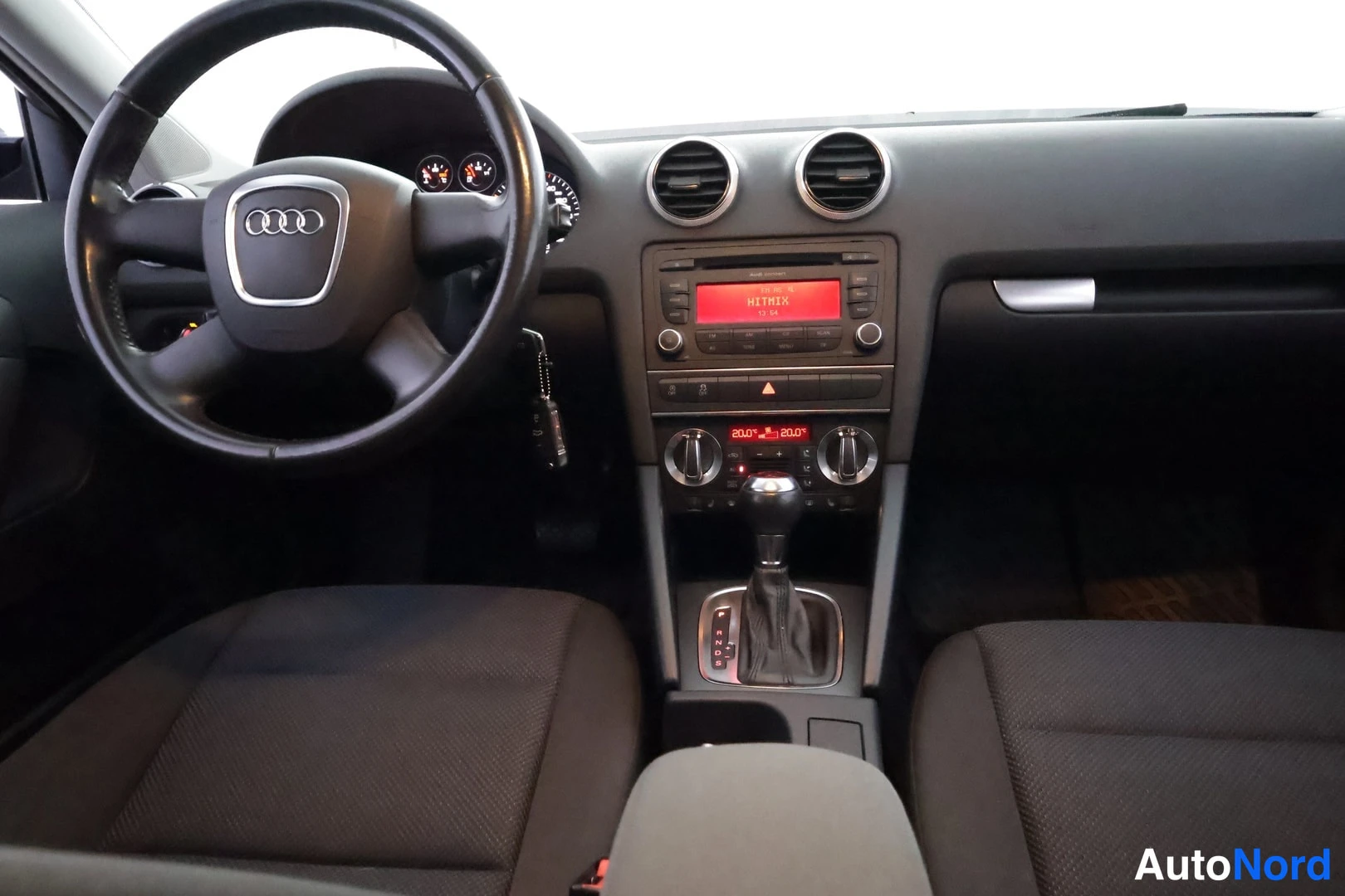 Muu: Sisäkuva ohjaamosta hieman sivulta: Audi-ohjauspyörä vasemmalla, mittarit, radio punaisella näytöllä, automaattivaihteiston valitsin ja tummat kankaiset etuistu