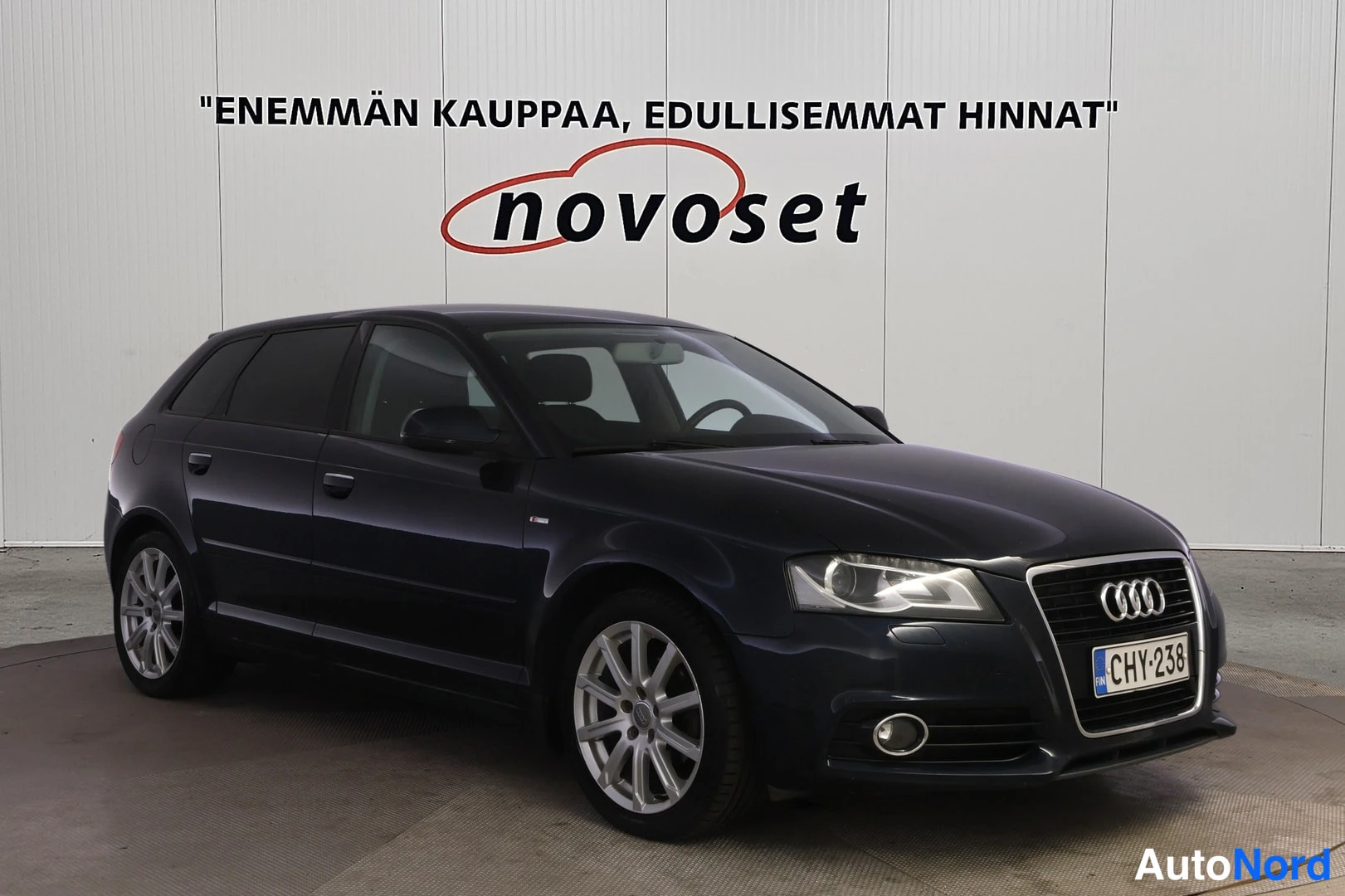 Etuviisto, nokka oikealle: Tumman sinertävän musta Audi A3 Sportback -farmari etukulmasta oikealta, rekisterillä, studion valkoista seinää vasten.