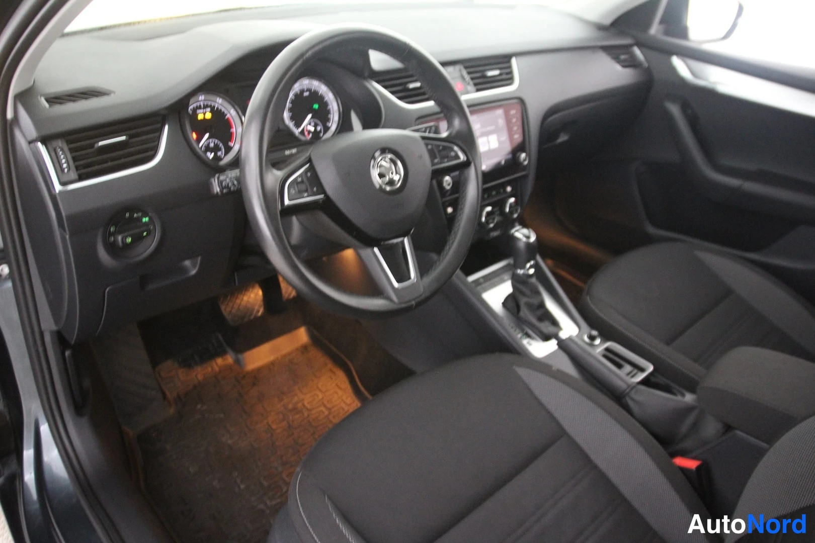 Muu: Ohjaamo kuljettajan puolelta: Skoda-ohjauspyörä, mittaristo varoitusvaloineen, keskikonsolin näyttö, automaattivaihteen valitsin ja mustat kankaankpoksuolat.