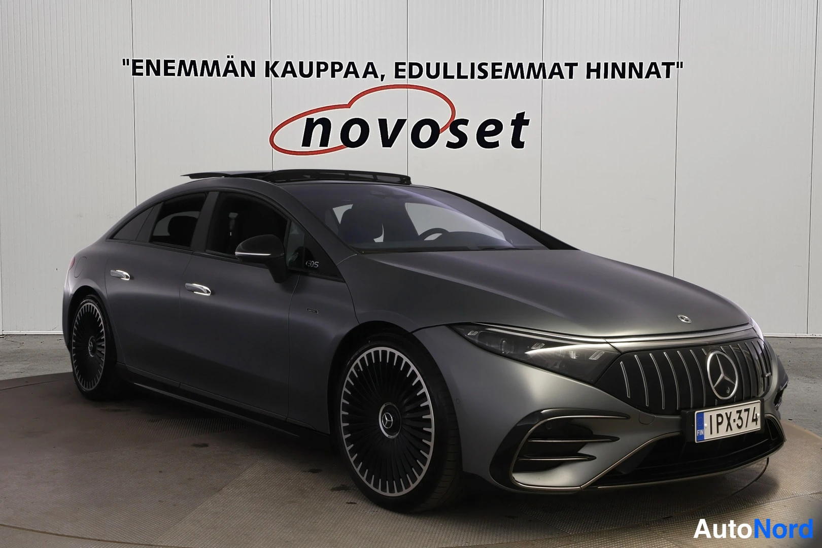 Etuviisto, nokka oikealle: Mercedes-Benzin etukulma oikealta, tummanharmaa sedan, taustalla valkoinen Novoset-seinäkyltti.
