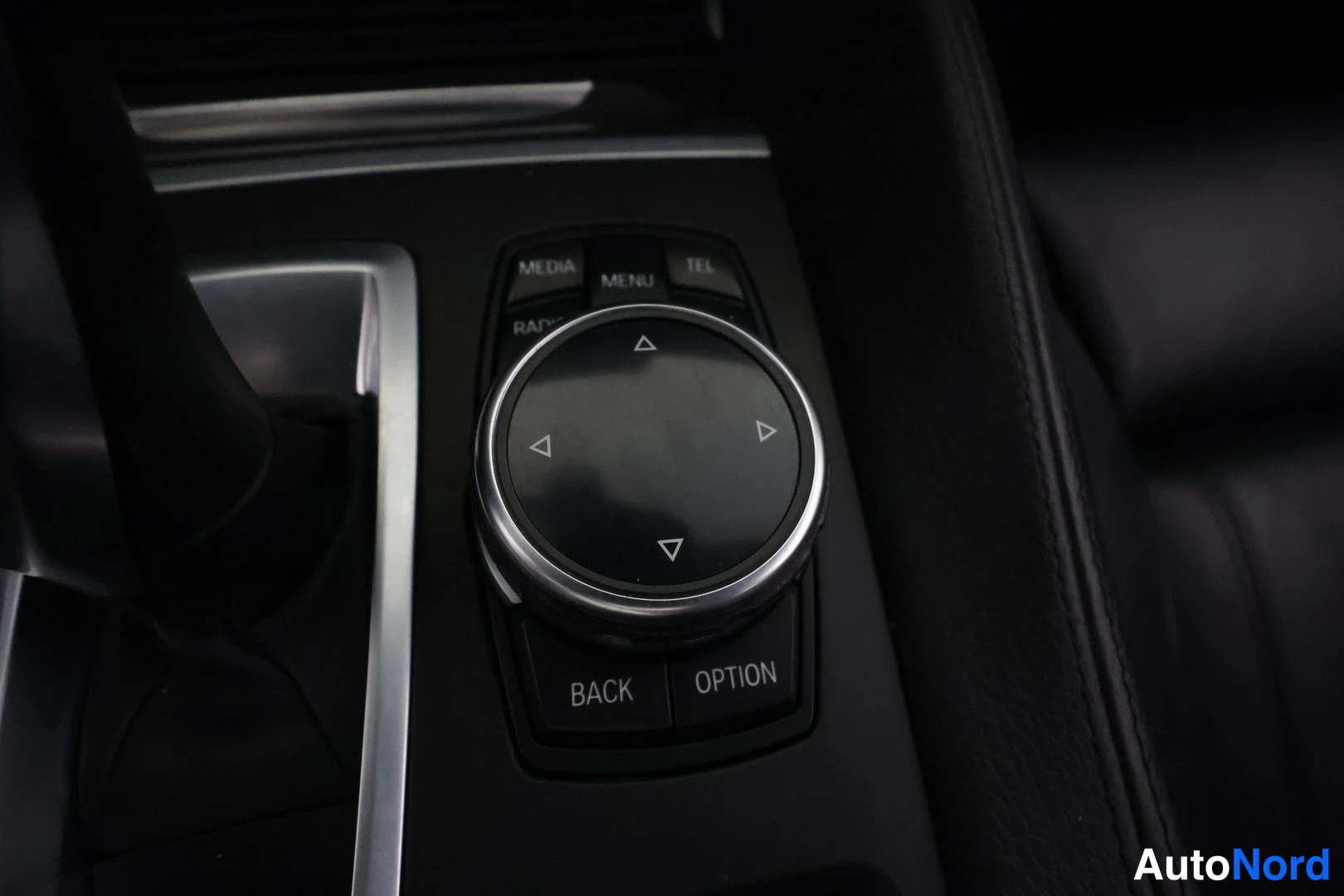 Muu: Lähikuva iDrive-ohjaimesta: pyöreä ohjainpainike MEDIA-, MENU-, TEL- ja RADIO-näppäinten keskellä, alla BACK- ja OPTION-painikkeet.