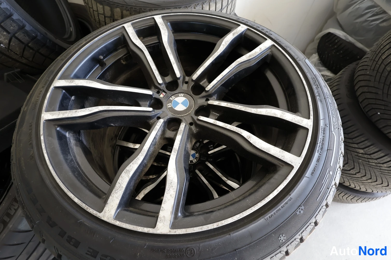 Yksityiskohta: Kuva renkaasta ja BMW-merkkisestä vanteesta varastoympäristössä, muiden renkaiden ympäröimänä.