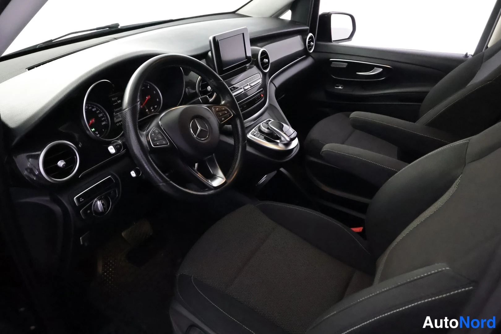 Ohjaamo: Driver-side interior shot of a dashboard and driver's seat. Steering wheel features multi-function buttons and a central logo. Center stack has an infotainment
