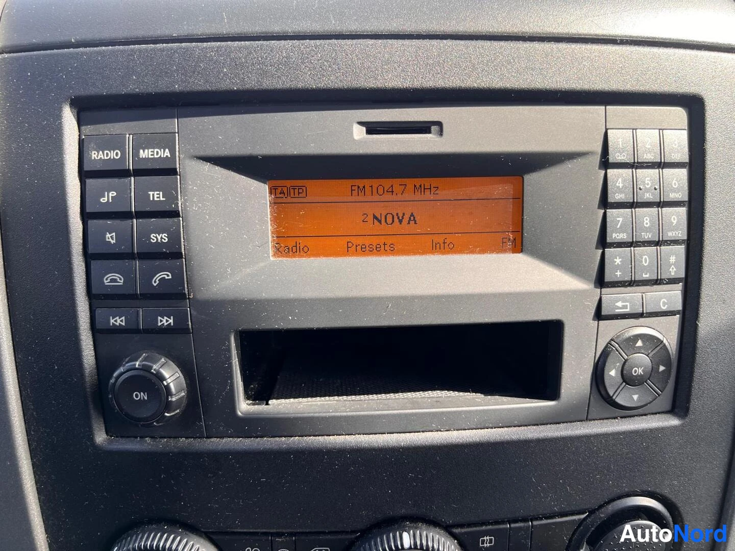 Yksityiskohta: Kojelaudan radioyksikkö lähikuvana: oranssi näyttö näyttää tekstin 'FM 104.7 MHz' ja 'NOVA', painikkeita RADIO, MEDIA, TEL, SYS sekä numeronäppäimistö ja OK-nup