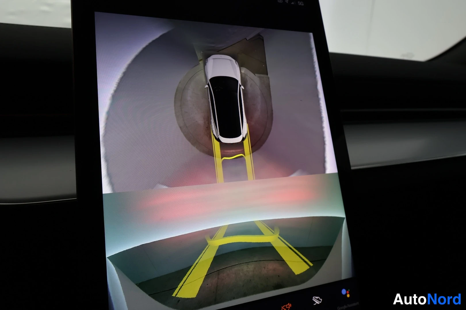 Muu: Auton infotainment-näytön valokuva. Näytöllä on ylhäältäpäin kuvattu kamera- tai tarkkailuohjelmanäkymä, jossa valkoinen auto seisoo pyöreällä tasanteella ja ke