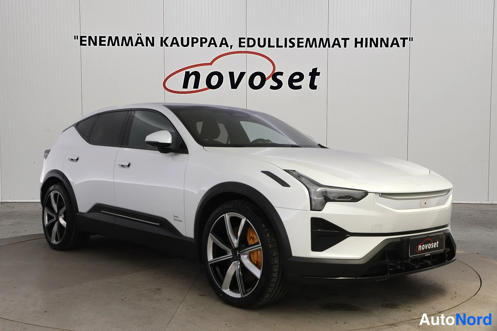 Etuviisto, nokka oikealle: Valkoinen Polestar 3 kulmaetunäkymästä, keula oikealla, näkyvät etugrilli, ajovalot, ovet ja mustat vanteilla varustetut pyörät sisätiloissa.