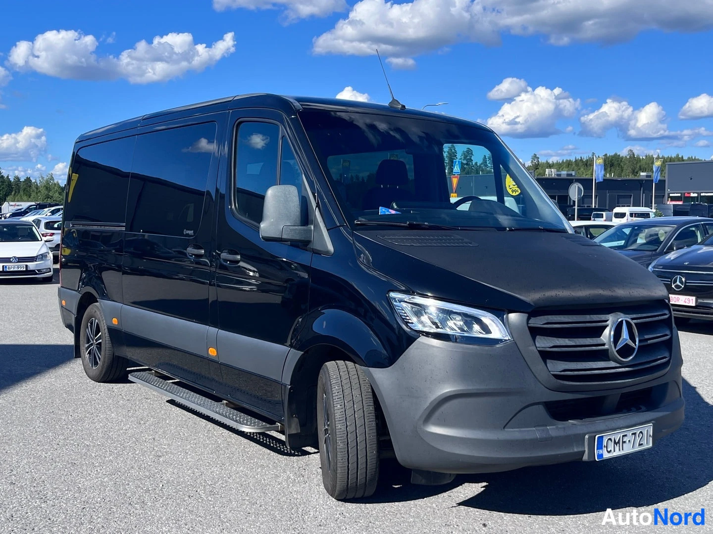 Etuviisto, nokka oikealle: Musta Mercedes-Benz Sprinter etuviistöstä oikealta; näkyvissä LED-ajovalot, astinlauta ja ovipeilit.