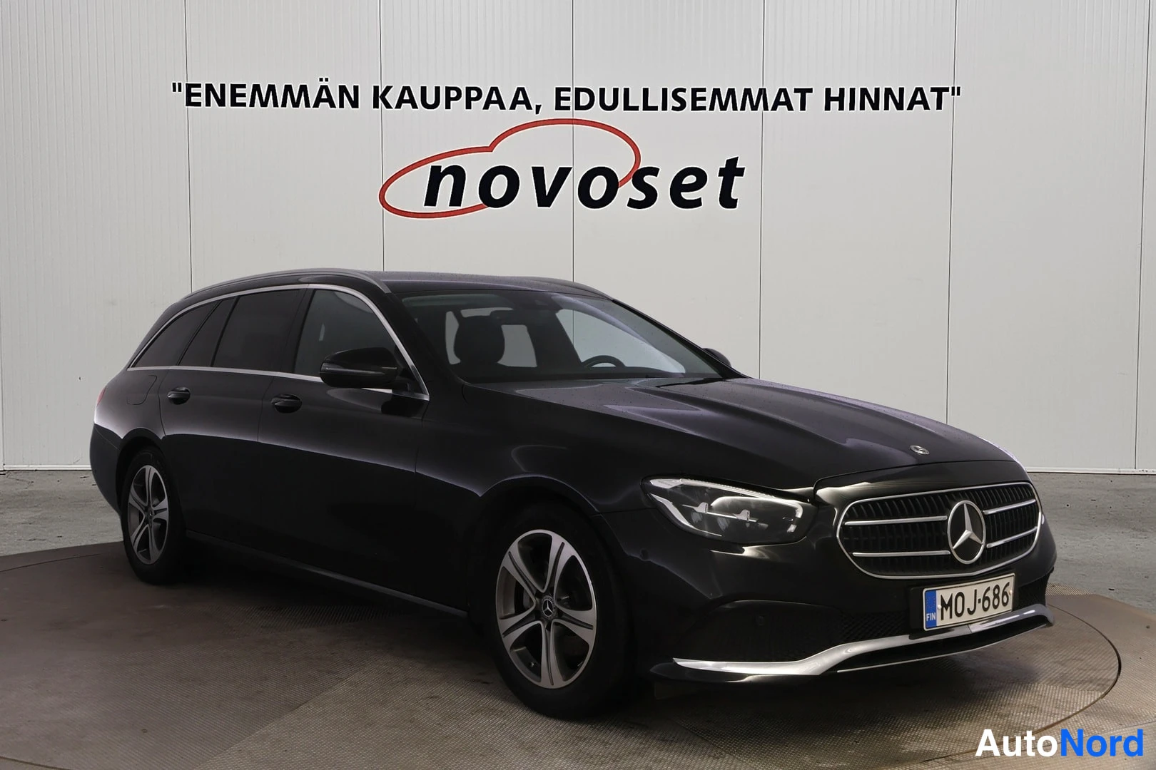 Etuviisto, nokka oikealle: Musta Mercedes-Benz E-mallin farmariauto etuviistokuva. Takalaatikko ja keulakuva näkyvät. Autossa on LED-ajovalot ja suuri merkki grillissä.