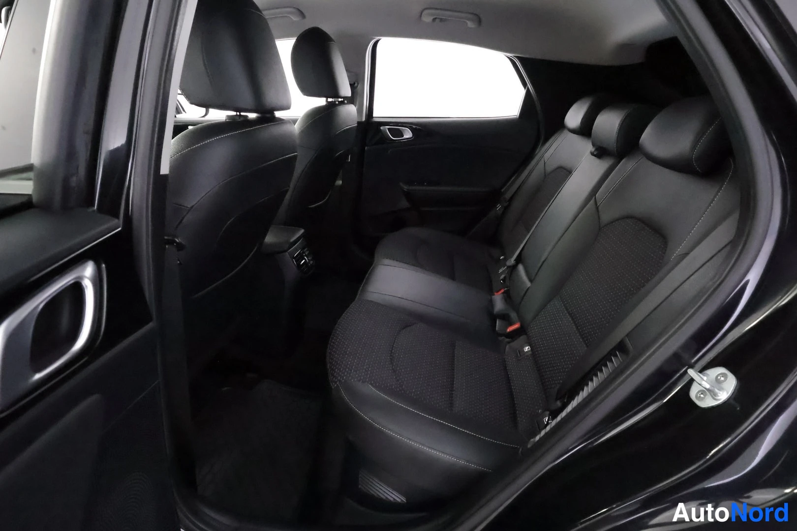 Takaistuimet: An open rear door reveals the second row of three black leather-like seats featuring white contrast stitching against a dark interior.