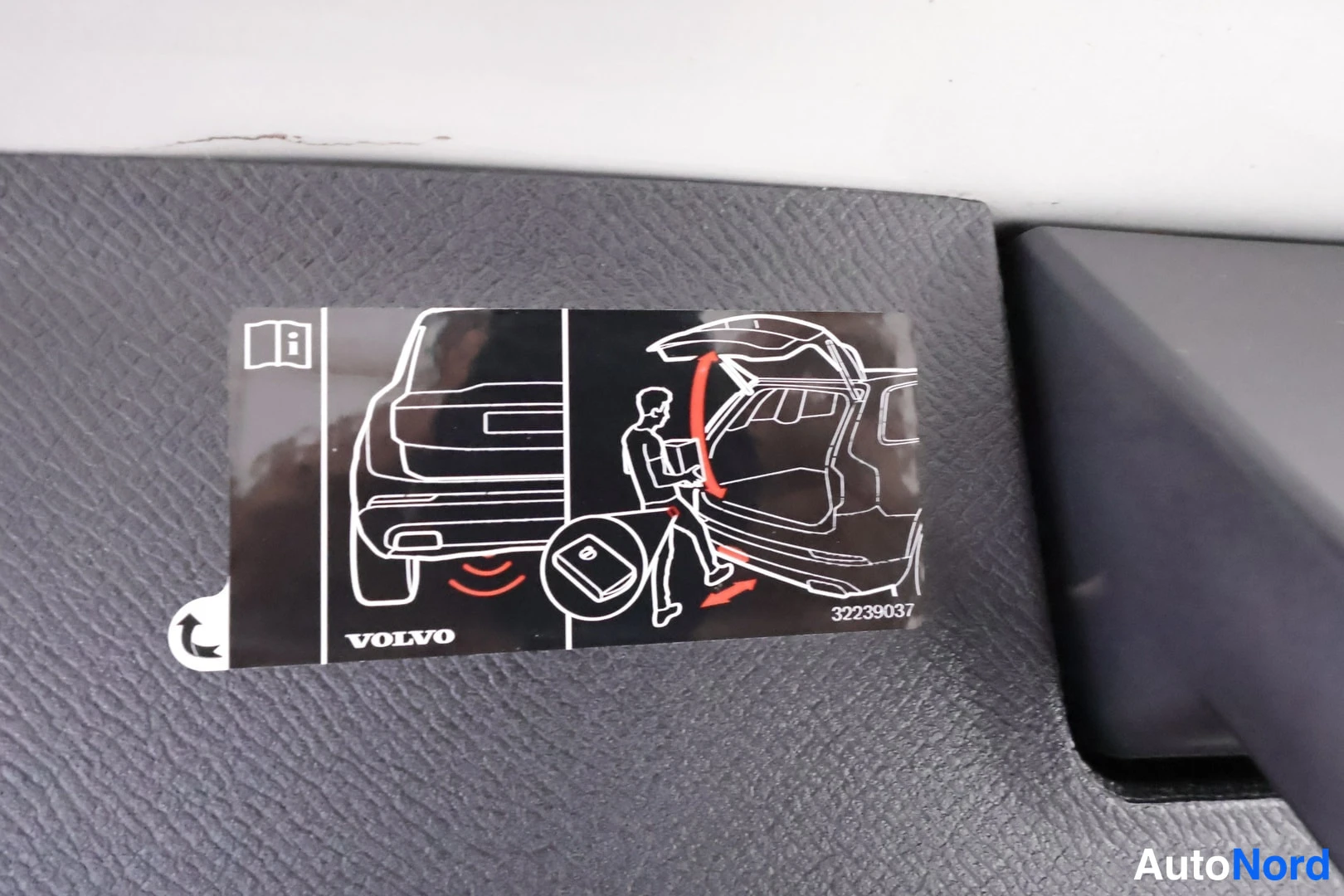 Yksityiskohta: Volvo-tarra tummassa muovipaneelissa, jossa kuvia sähkötoimisen takaluukun avaamisesta ja latauspataluvusta, teksti VOLVO ja numero 32239037.