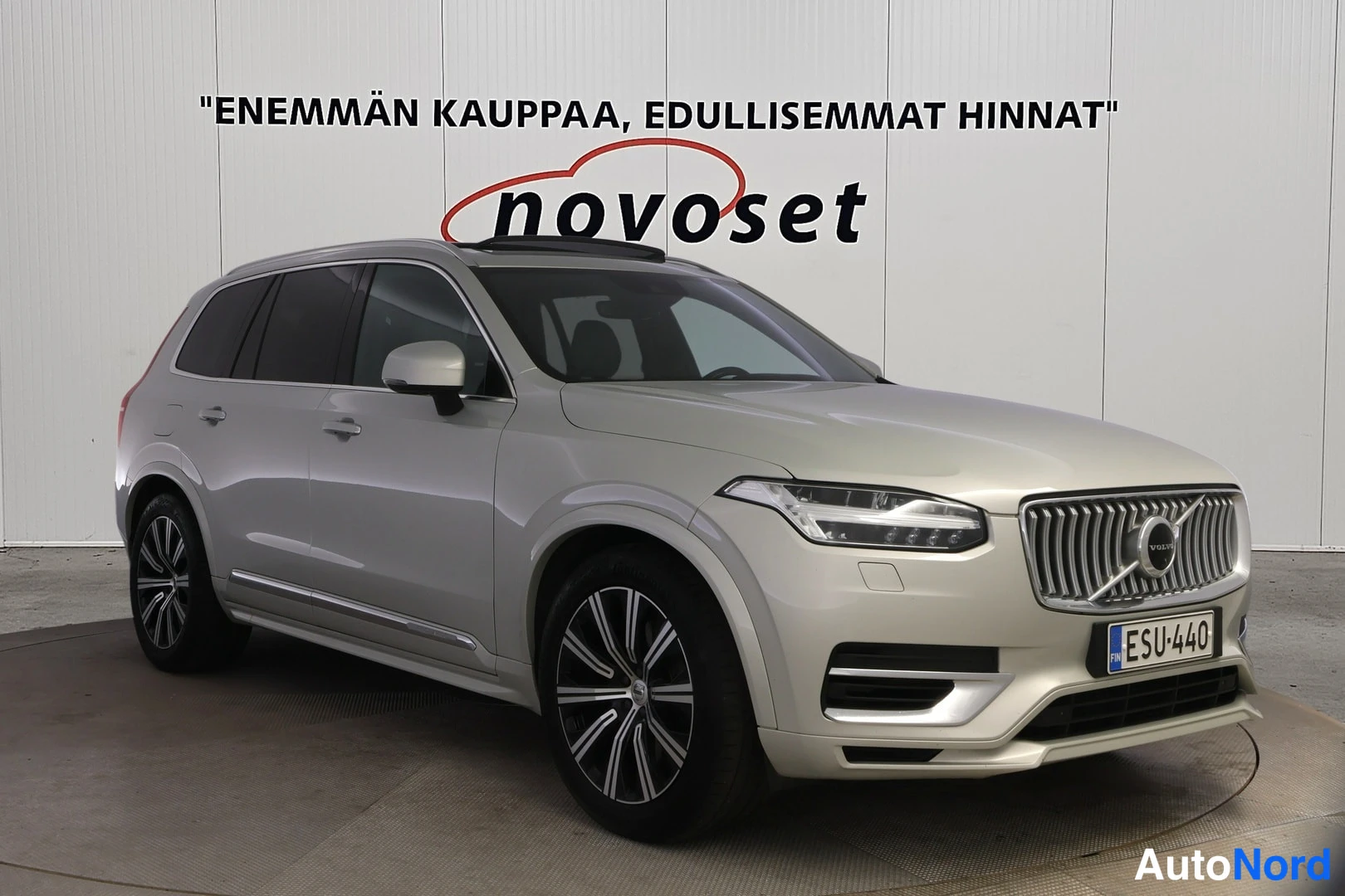 Etuviisto, nokka oikealle: Etukolmaisessa kuvassa vaaleanbeige Volvo XC90 -maasturi: etusäleikkö ja ajovalot, lasinen panoraamakatto, kattokaiteet ja rekisteri.