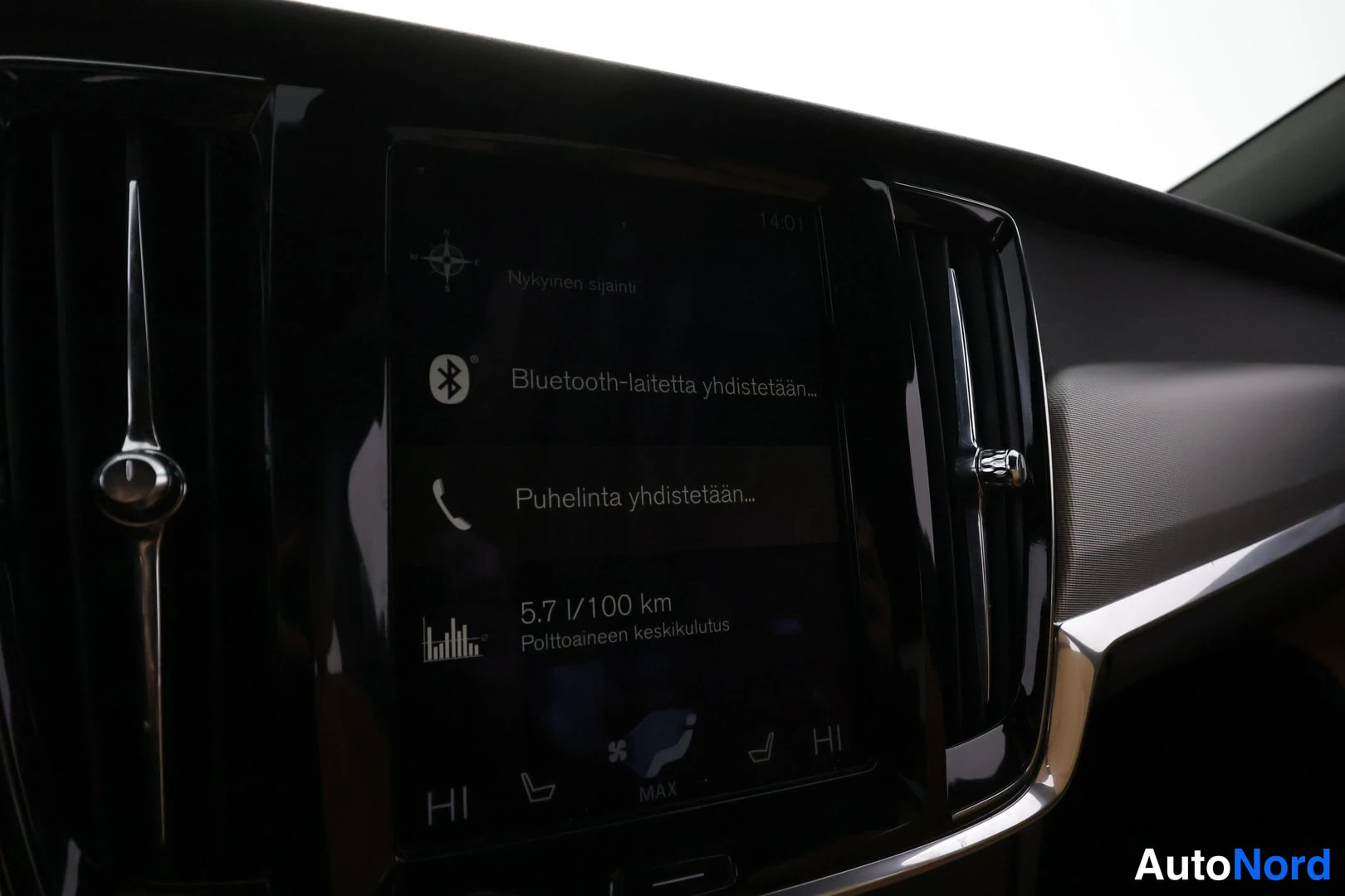 Ohjaamo: Close-up of the car's vertical centre touchscreen. The display shows Bluetooth connecting status and trip computer consumption in Finnish.