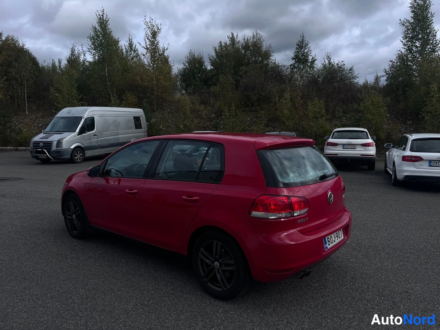 Takaviisto: Punaisen Volkswagen Golfin takasivu nähtynä kauempaa, osittain varjossa. Auto seisoo parkkipaikalla metsän reunalla. Takaosa ja oikea kylki näkyvät. Takavalot p