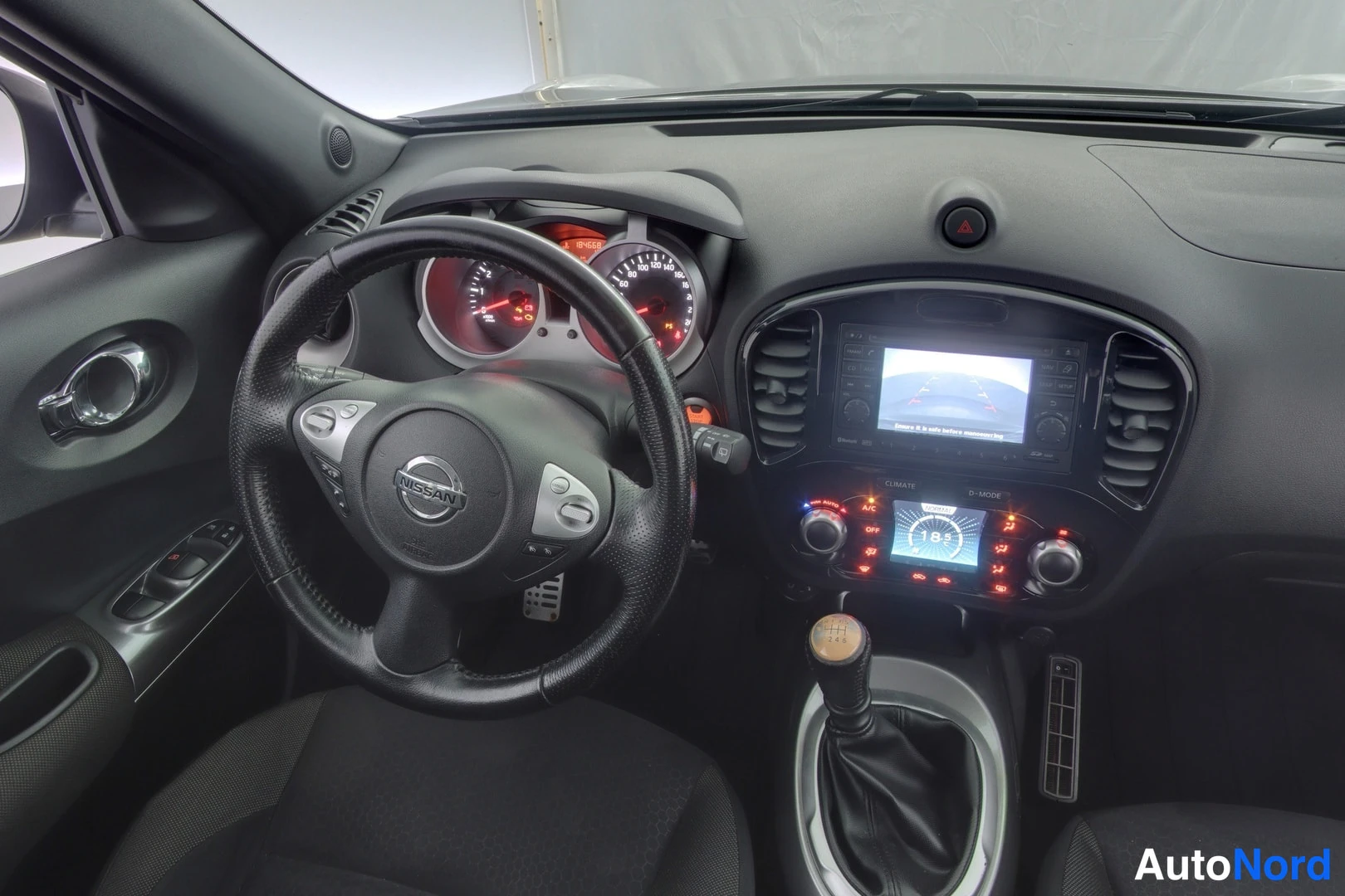 Edestä: Nissan Juke kojelauta lähempää, mittaristo valaistuna, keskinäyttö ja manuaalivaihto