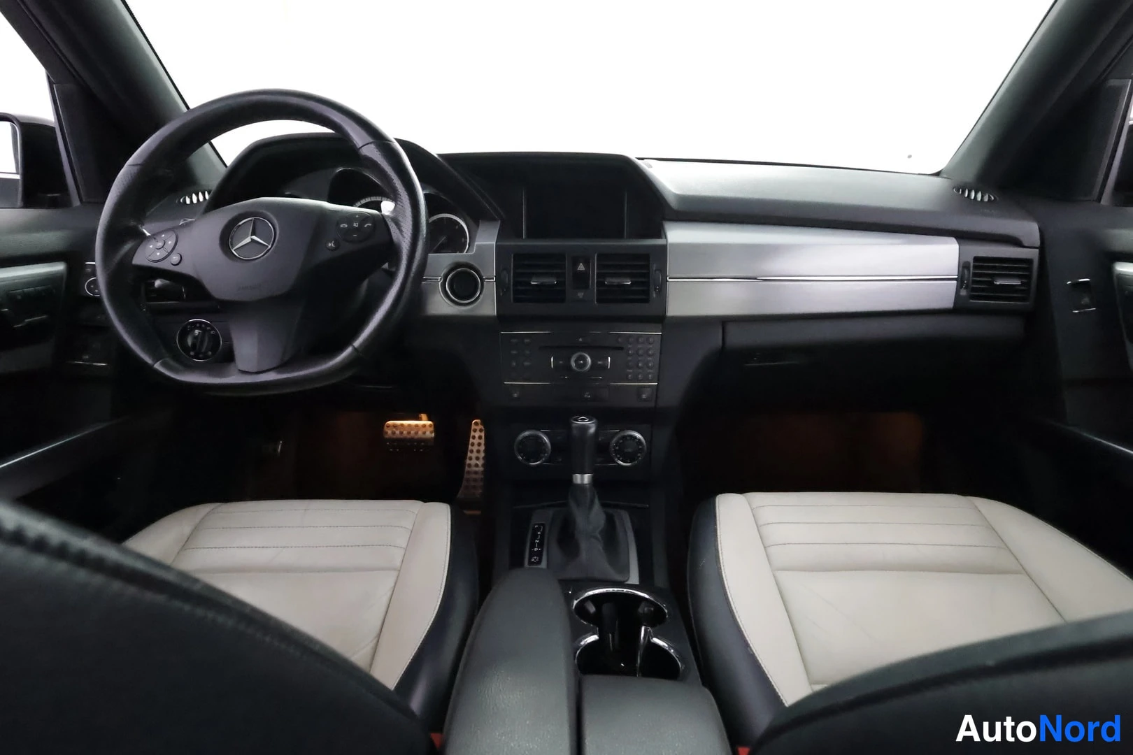Muu: Ohjaamon etunäkymä: Mercedes-Benz-merkitty ohjauspyörä, tumma kojelauta hopeisella paneelilla, mittarit, radioyksikkö ja vaaleat etuistuimet.