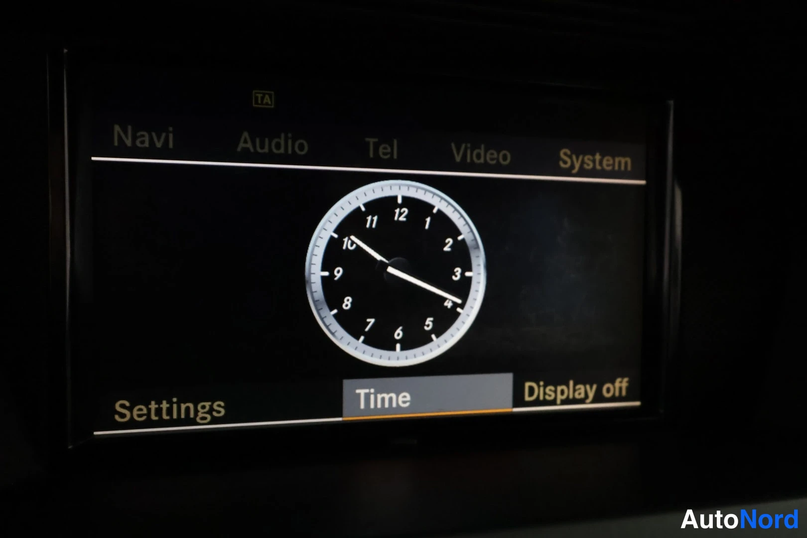 Yksityiskohta: Auton kojelaudan infotainment-näyttö, jossa analoginen kello ja valikko: Navi, Audio, Tel, Video, System, Settings, Time, Display off.