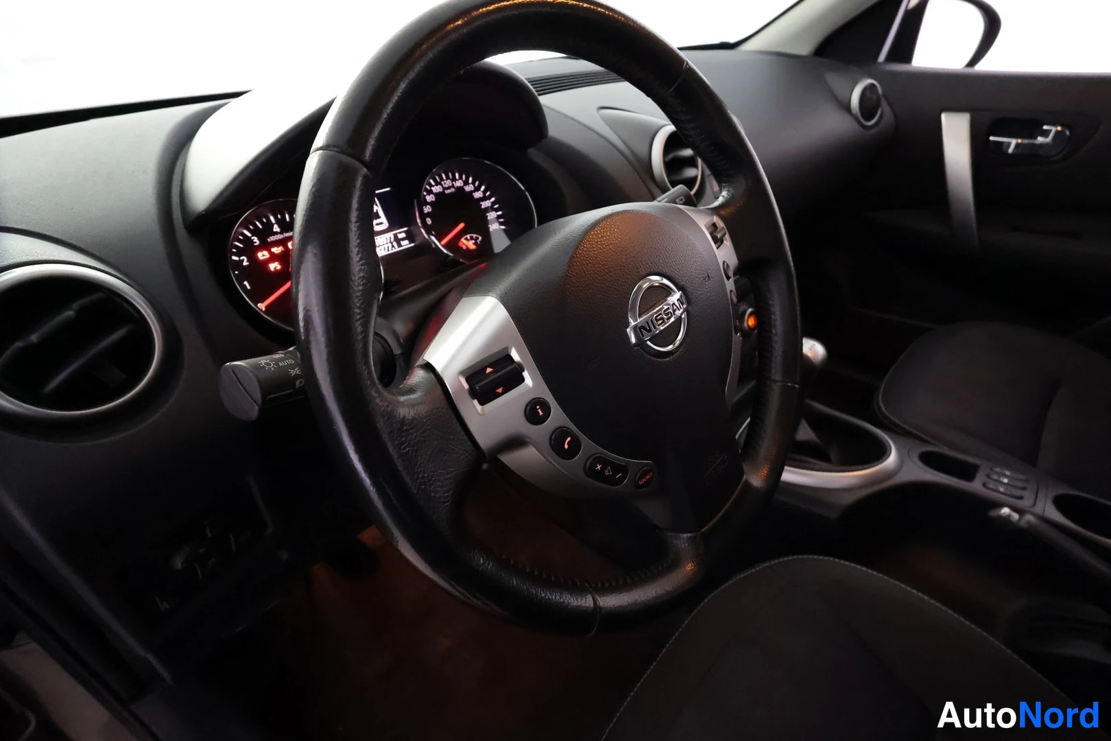 Edestä: Ratin ja kojelaudan tarkempi kuva; Nissan-merkki ratissa, mittaristö valaistu.